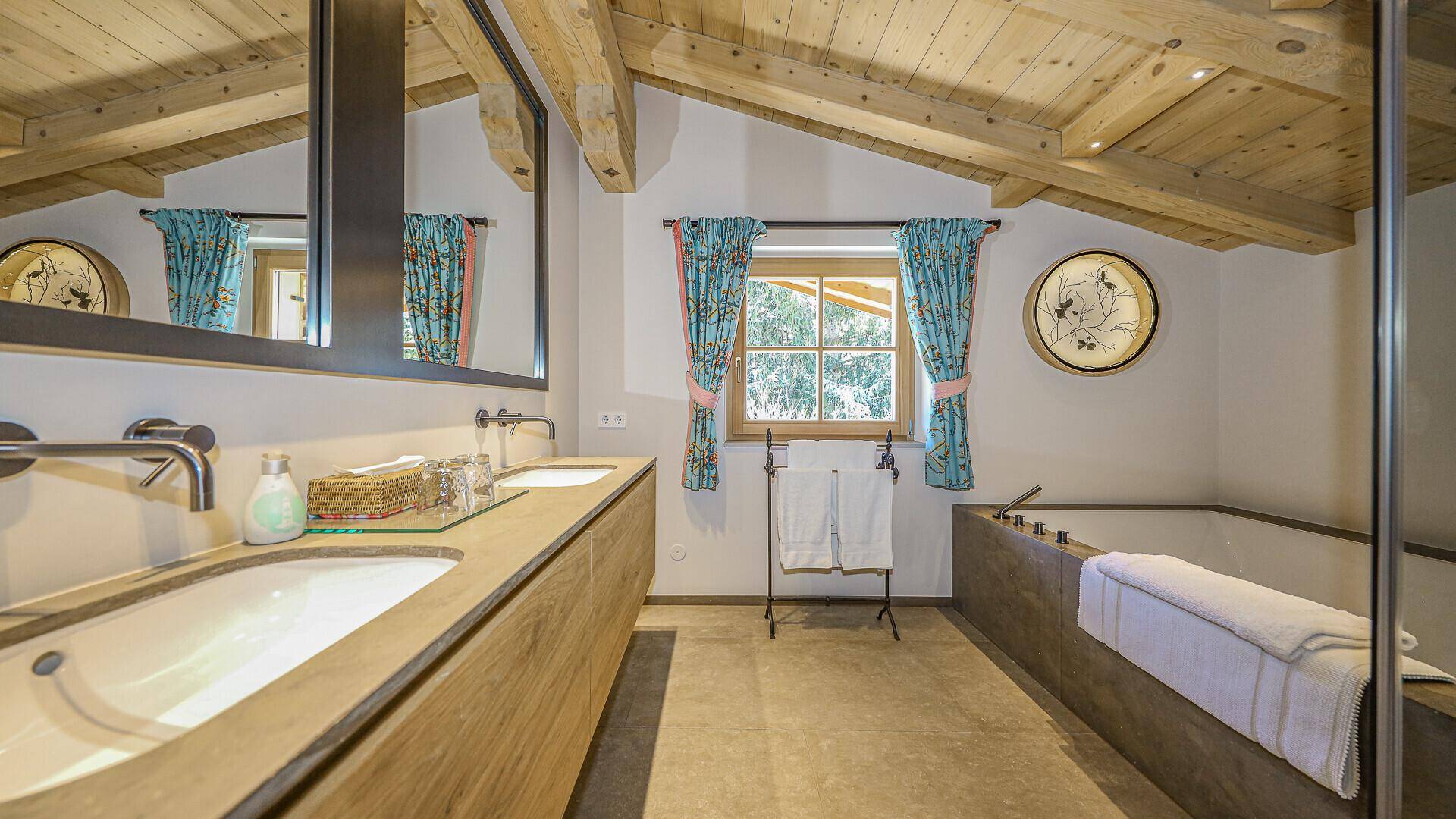 Helles Badezimmer mit Doppelwaschbecken, Badewanne und Holzbalkendecke im alpinen Stil.