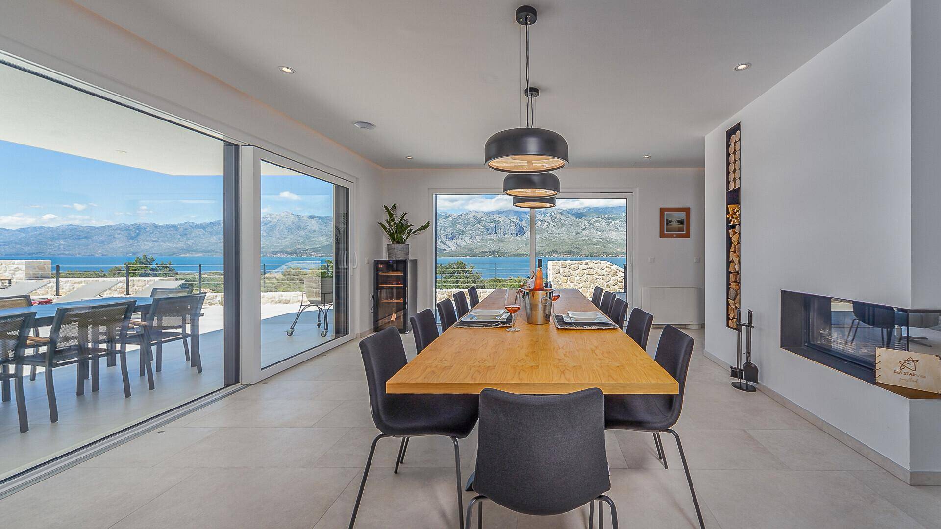 Eleganter Essbereich mit großem Holztisch, modernen Pendelleuchten und direktem Zugang zur Terrasse mit Meerblick.
