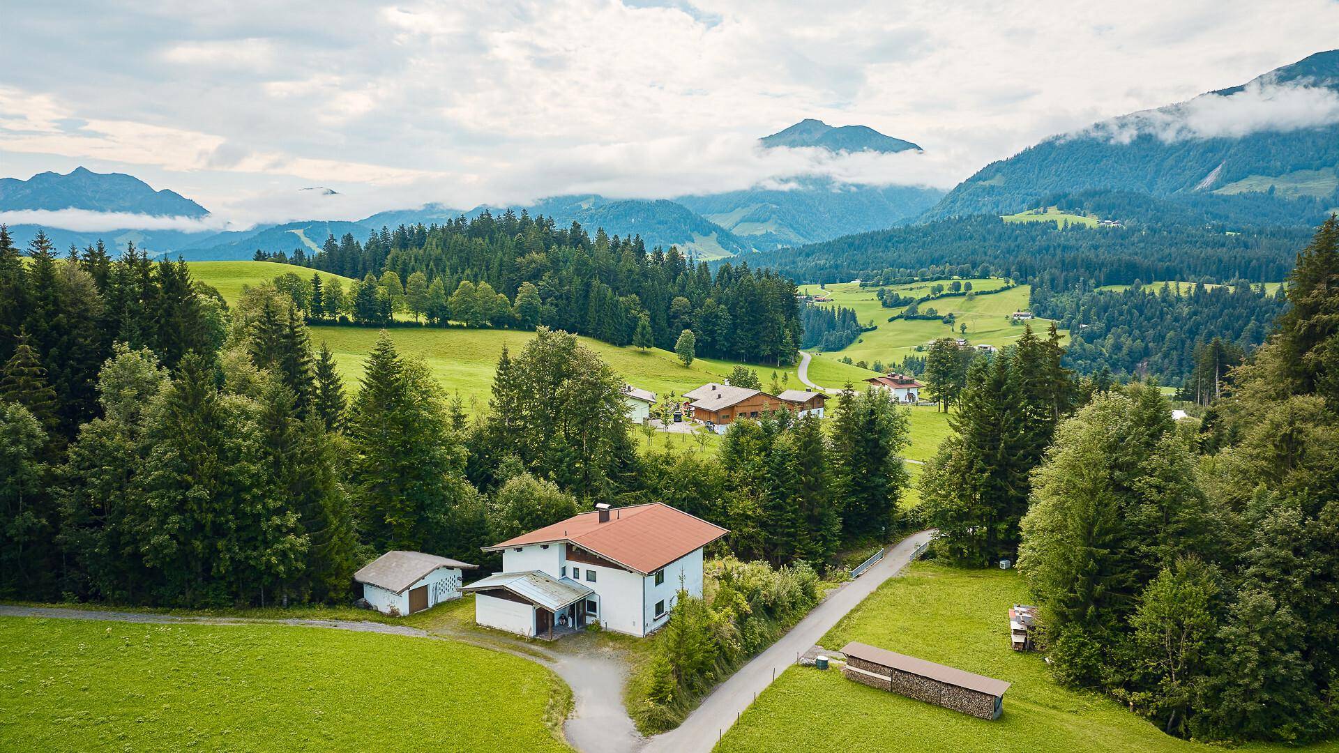 Luftaufnahme eines traditionellen Hauses mit rotem Dach in einer idyllischen grünen Hügellandschaft.