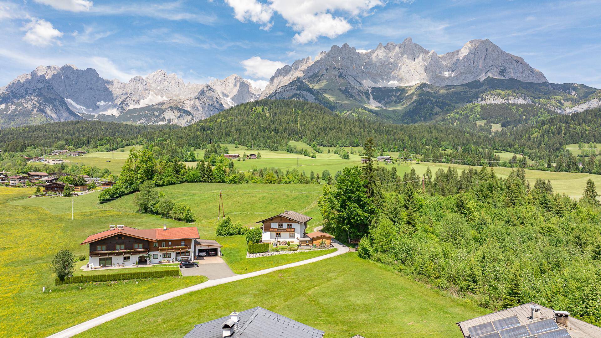 Weitläufige Landschaft mit mehreren Häusern und majestätischen Bergen im Hintergrund unter blauem Himmel.
