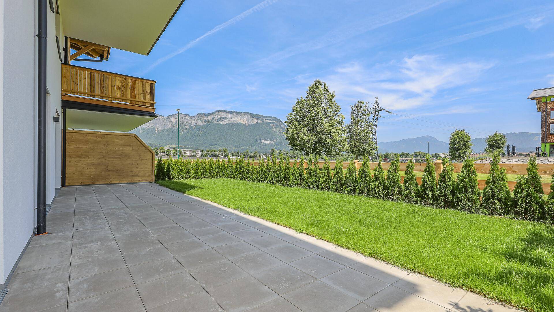 Große, gepflasterte Terrasse mit Rasenfläche und Hecke, umgeben von Bergen.