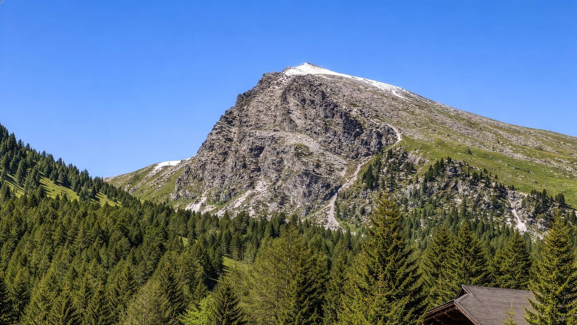 Ein majestätischer, felsiger Berggipfel mit spärlicher Vegetation und Schneeresten unter blauem Himmel.