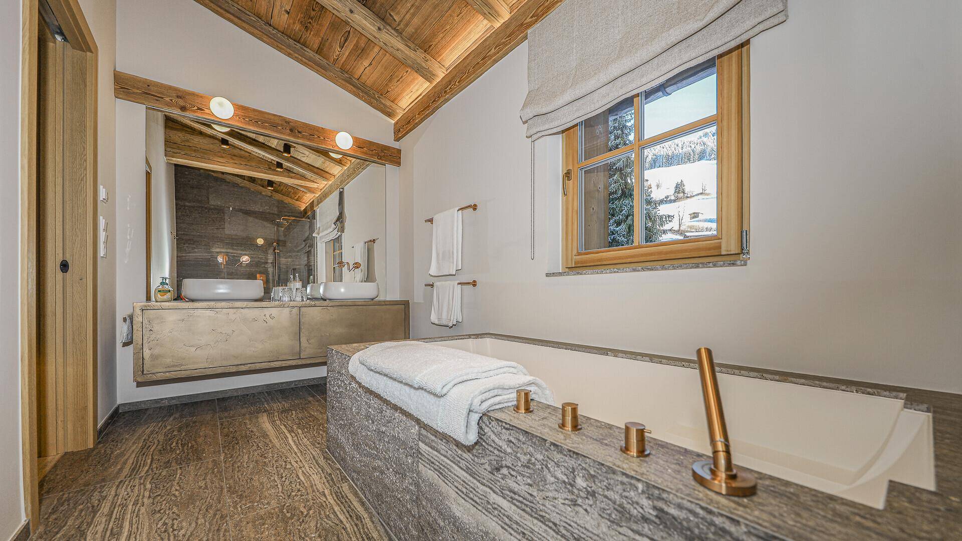 Elegantes Badezimmer mit Badewanne, Doppelwaschbecken und Holzbalkendecke im alpinen Design.