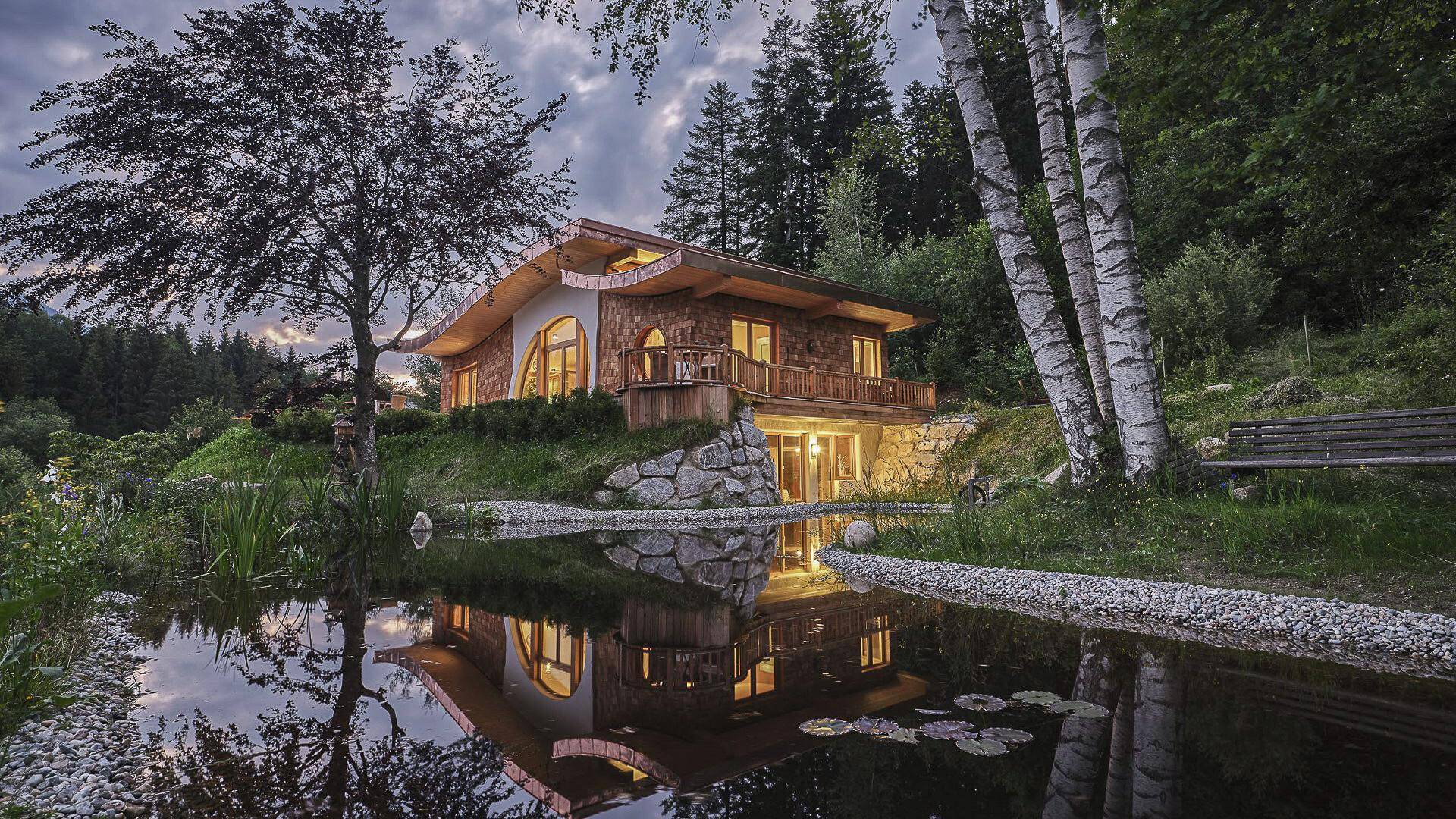 Traditionelles Alpenhaus bei Dämmerung, malerisch in einem Teich gespiegelt, umgeben von Bäumen.