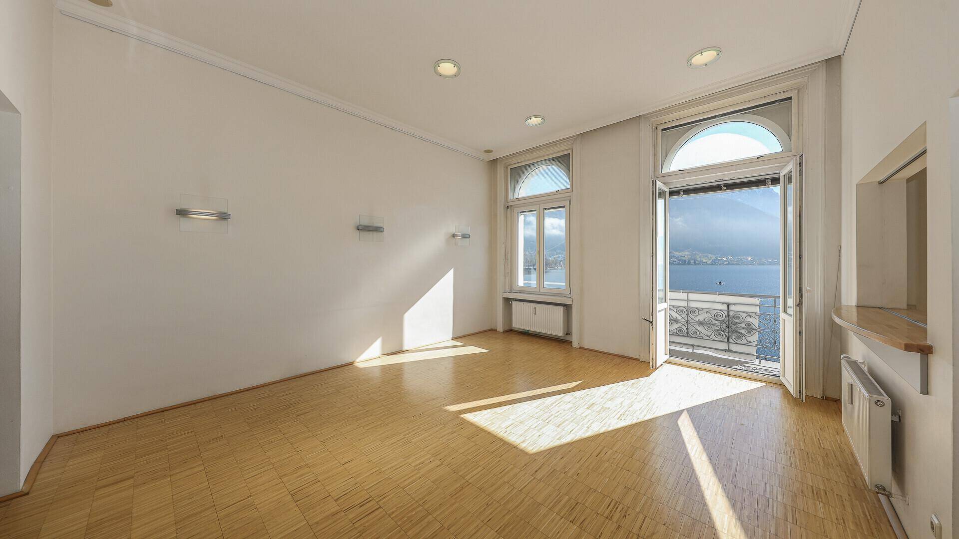 Lichtdurchfluteter leerer Wohnraum mit Parkettboden und direktem Zugang zum Balkon mit Seeblick.
