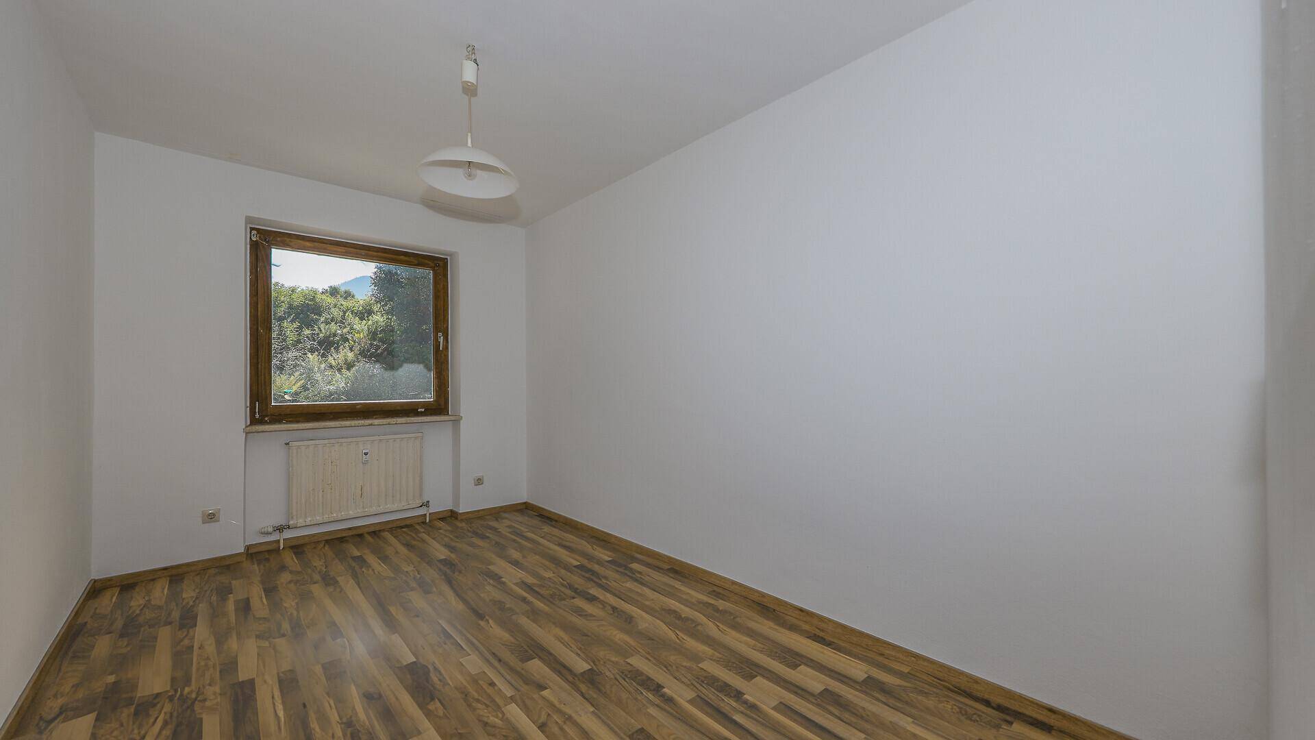 Leeres Schlafzimmer mit Holzboden, weißen Wänden, einem Fenster und Heizkörper.