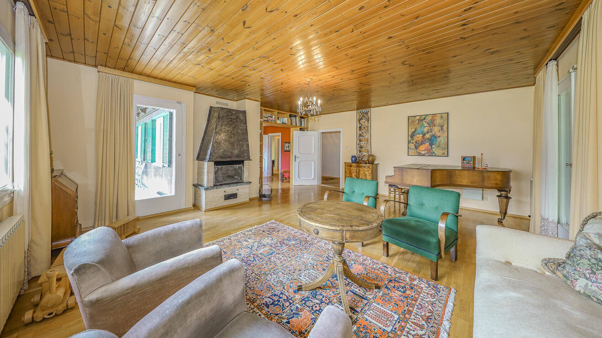 Weitläufiges Wohnzimmer mit Kamin, Holzdecke, Parkettboden und Zugang zum Balkon.