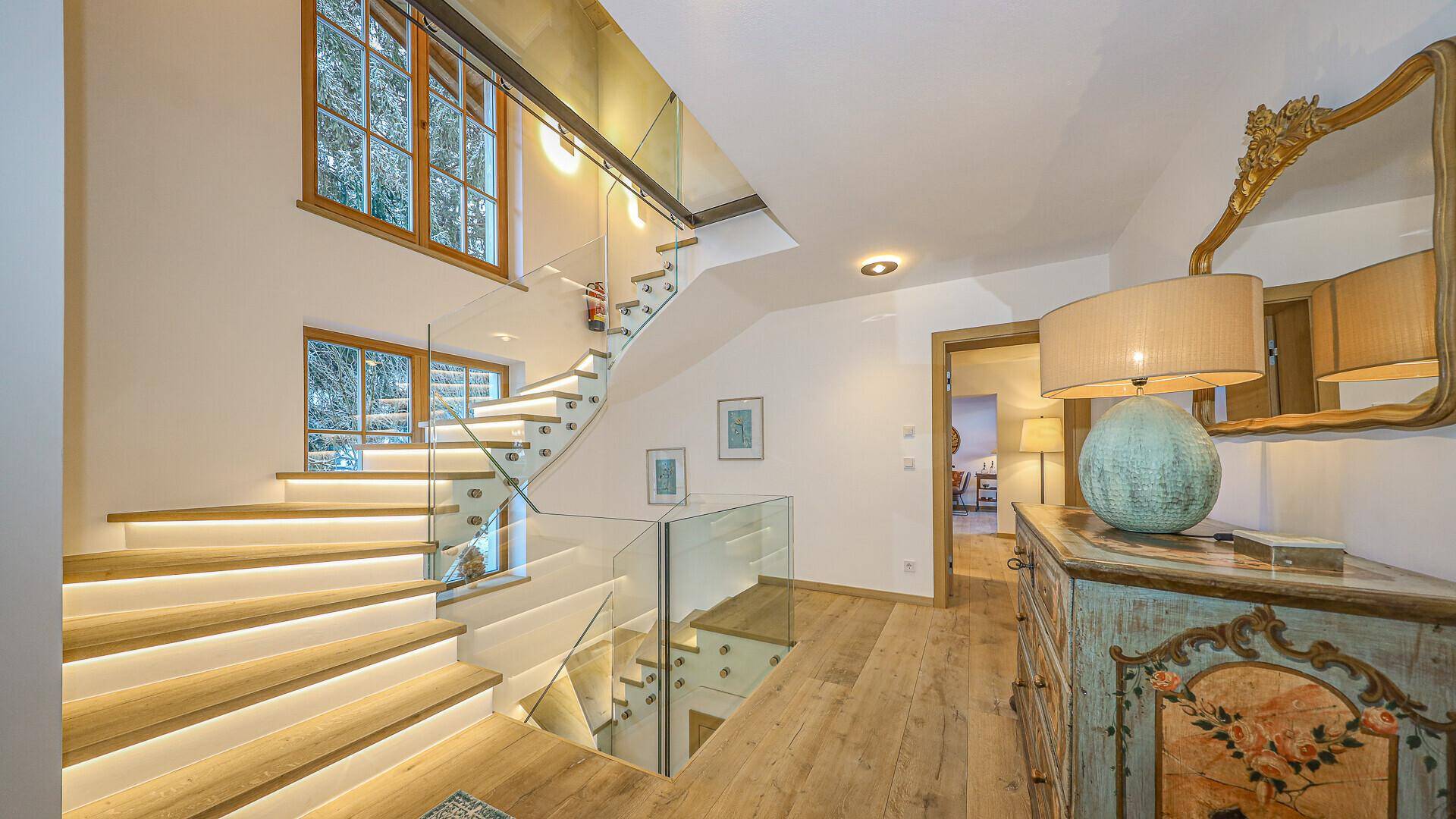 Heller Flur mit moderner Treppe, Glasgeländer und traditioneller Kommode im alpinen Stil.