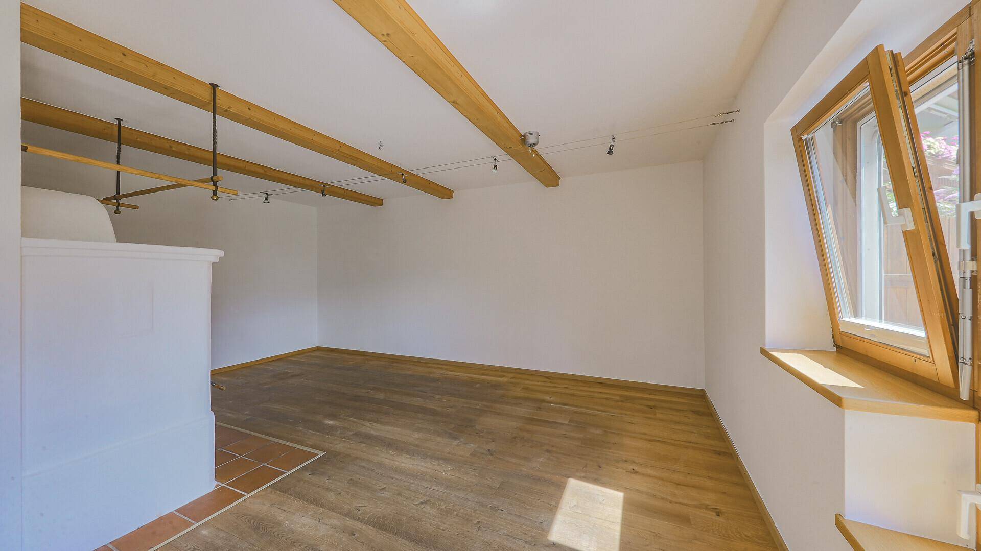 Heller Raum mit Holzboden, sichtbaren Holzbalken und einem traditionellen Kachelofen.