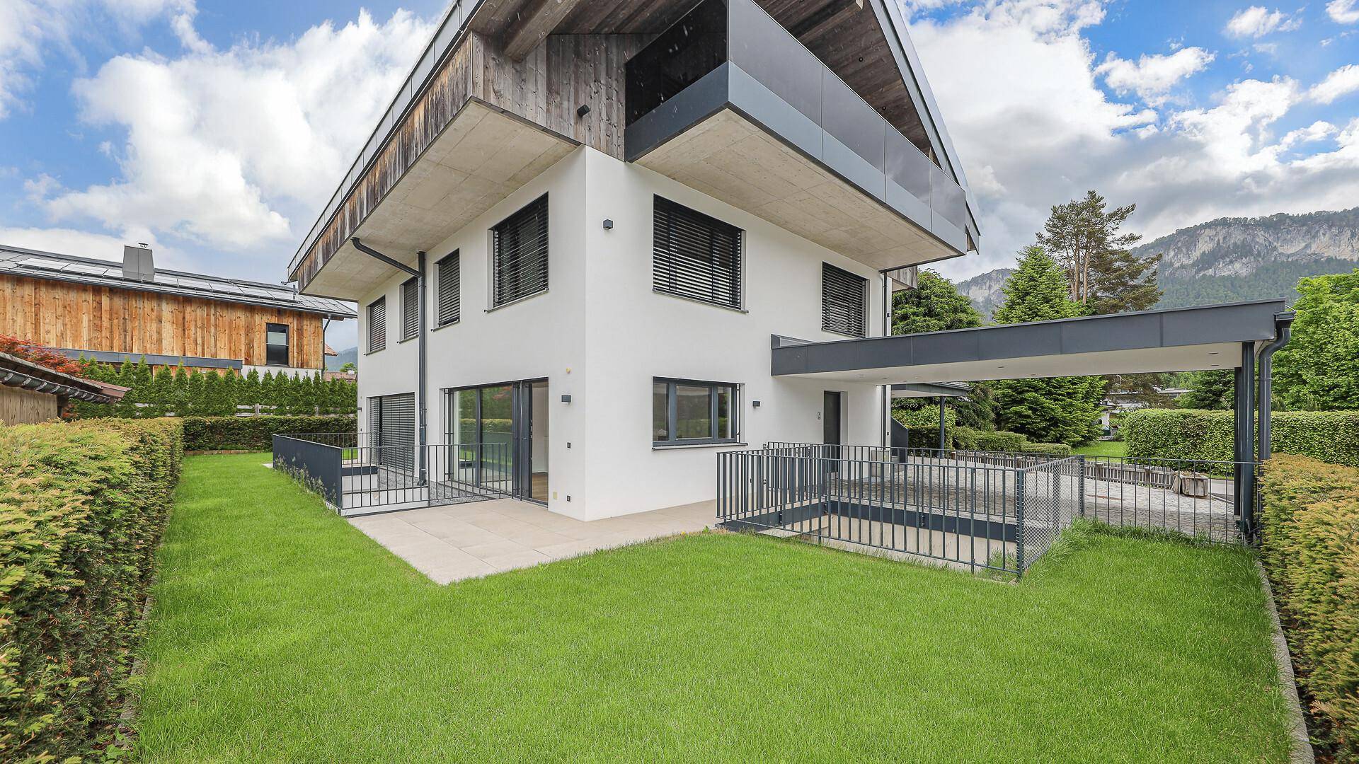 Modernes Haus mit gepflegtem Rasen, Terrasse und Bergblick unter blauem Himmel.