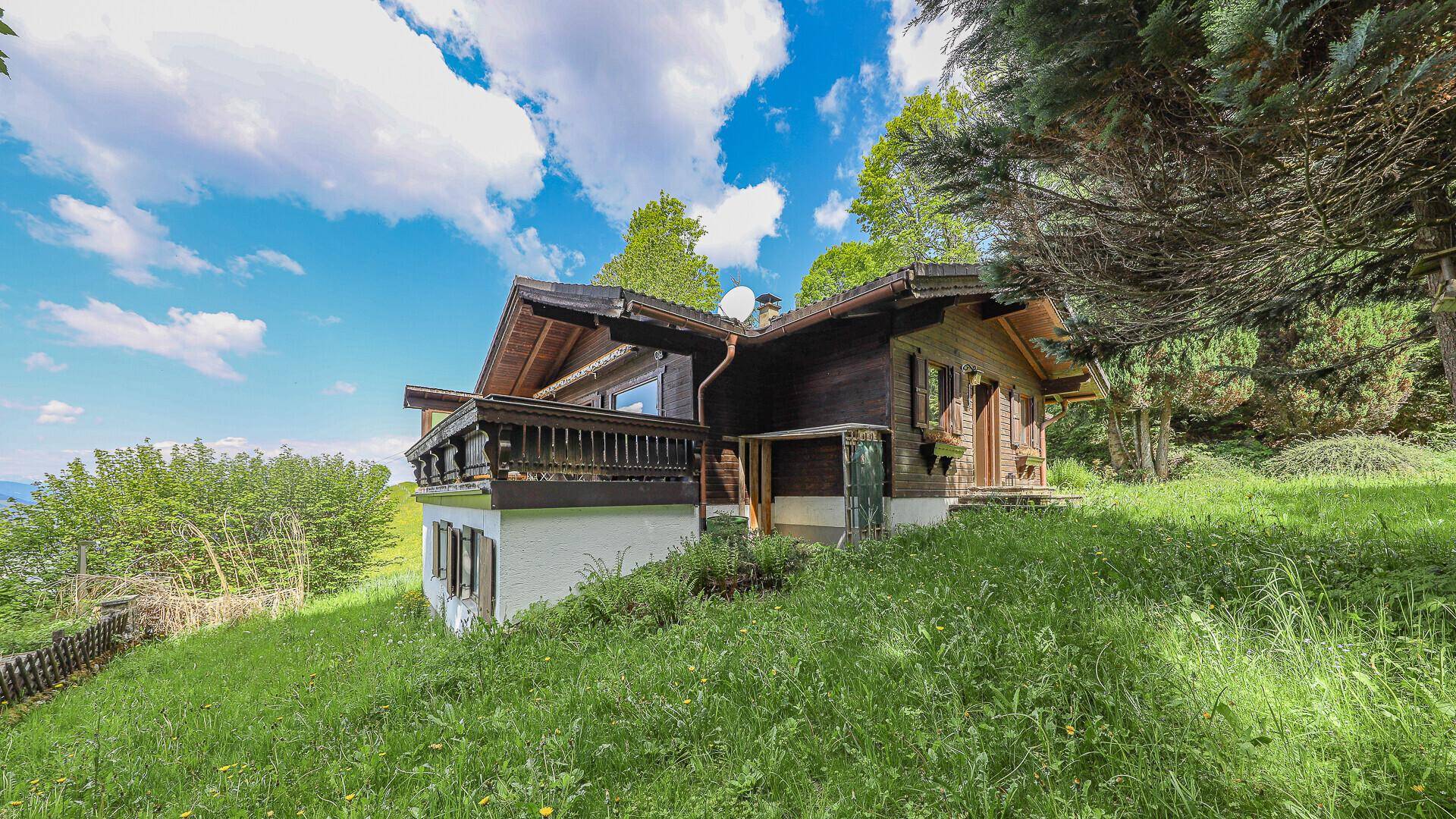 Traditionelles Holzchalet mit Balkon und Steinsockel, umgeben von grüner Natur und Bäumen.