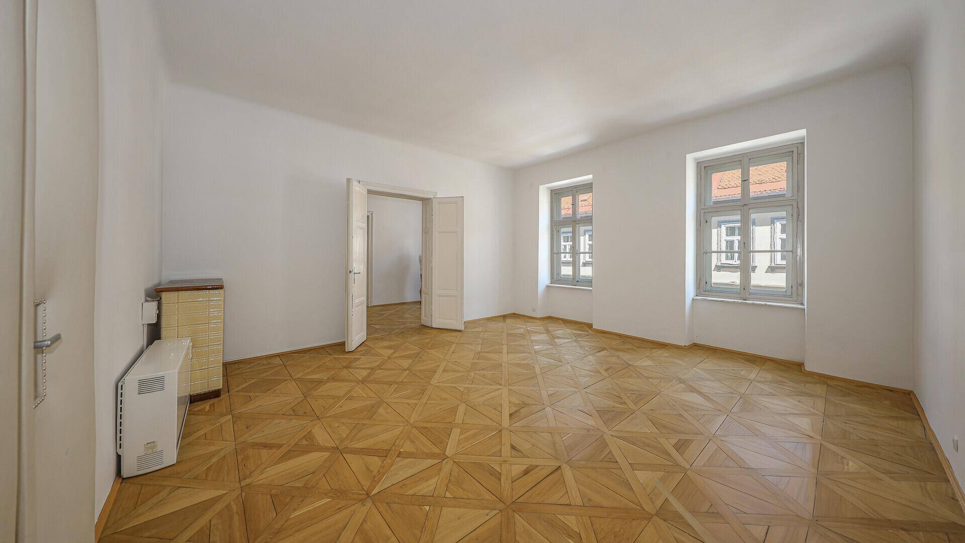 Großes, leeres Zimmer mit Parkettboden, Heizkörper und Blick auf die Straße.