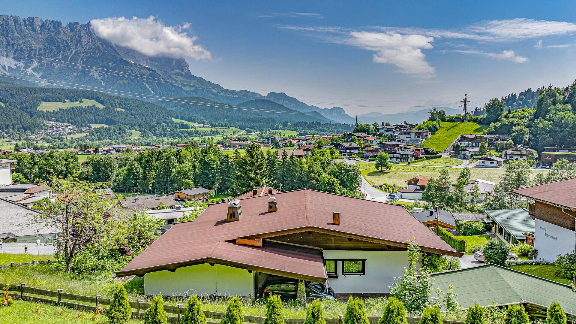 Traditionelles Haus mit braunem Dach und grüner Hecke vor beeindruckendem Bergpanorama.
