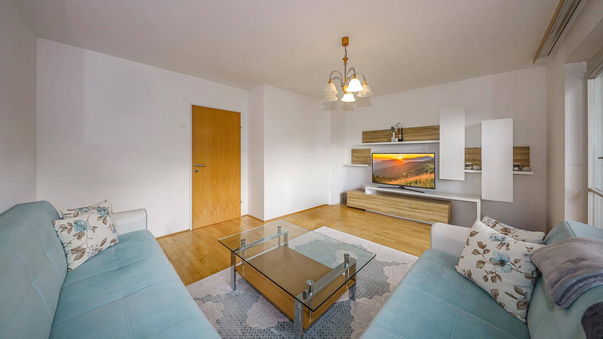 Helles Wohnzimmer mit türkisem Sofa, Holzboden und modernem TV-Möbel, mit Zugang zu einem Flur.