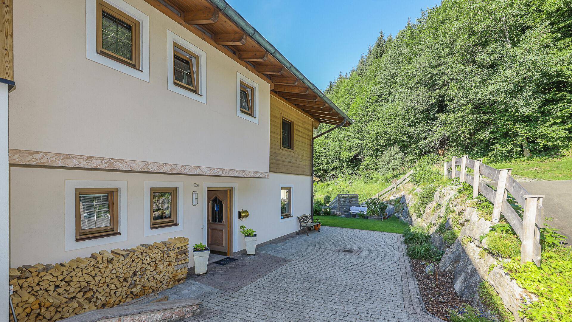 Eingangsbereich des Hauses mit gepflasterter Auffahrt, Holzstapel und Blick auf die grüne Umgebung.