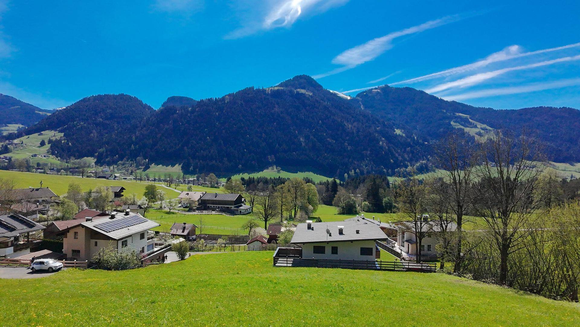 Weitläufiger Blick über eine ländliche Siedlung mit grünen Wiesen und bewaldeten Bergen.