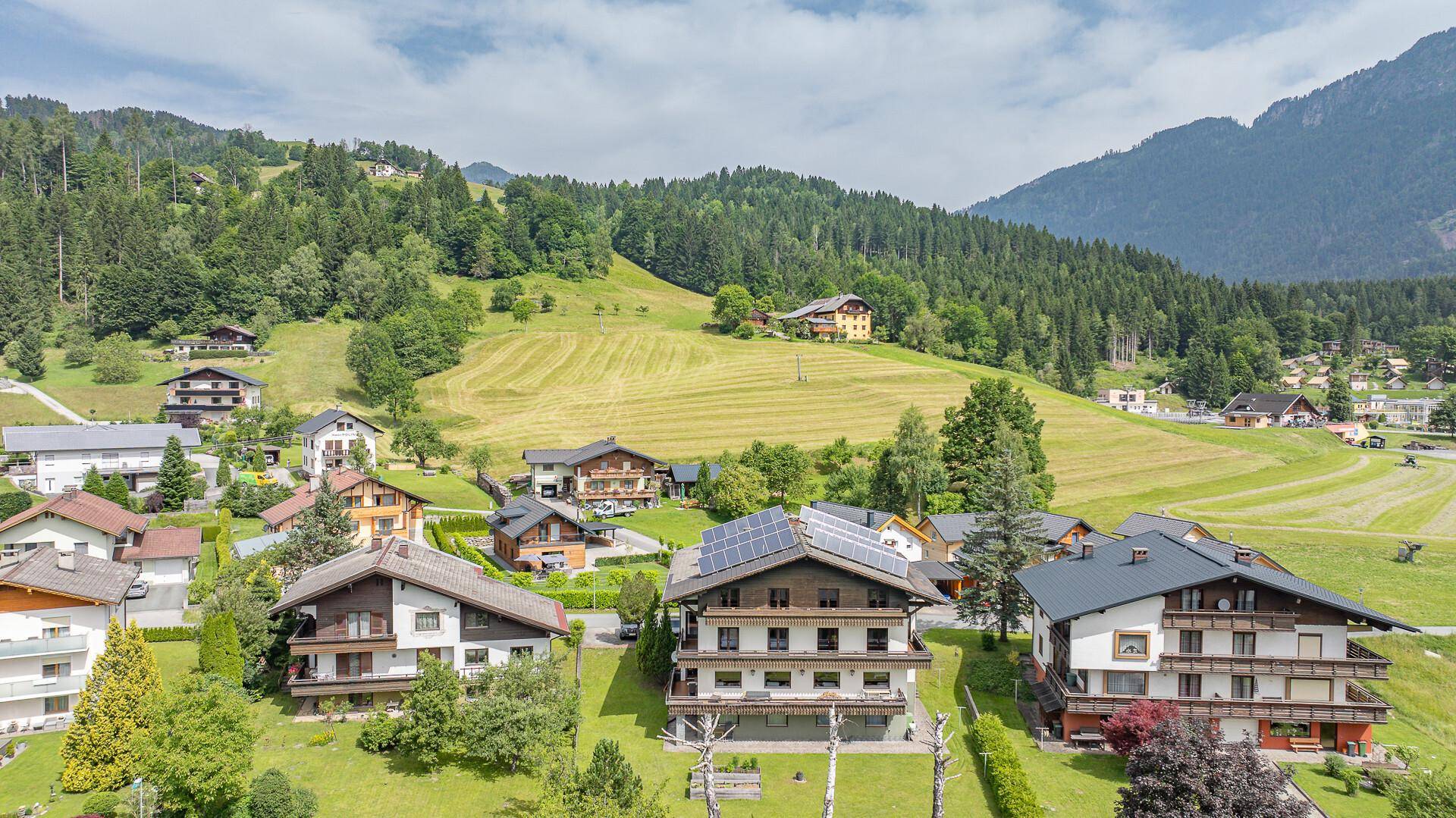 Weitläufige Luftaufnahme eines Dorfes eingebettet in eine grüne Hügellandschaft mit majestätischen Bergen im Hintergrund.