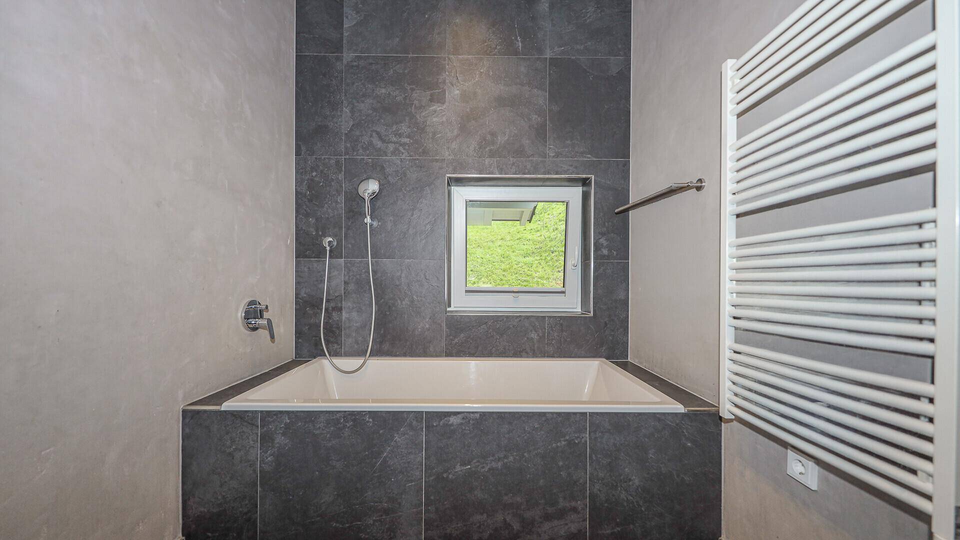 Elegante Badewanne in modern gefliestem Badezimmer mit Fenster und Handtuchheizkörper.