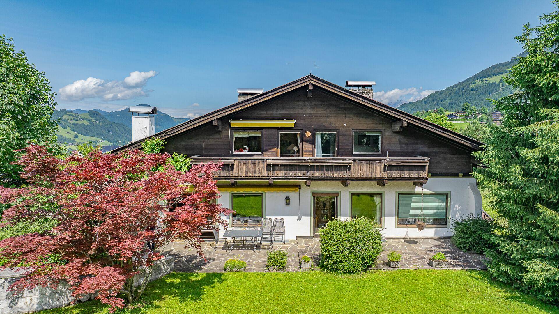 Rückseite des Chalets mit großer Steinterrasse, Gartenmöbeln und Blick auf die Berge.