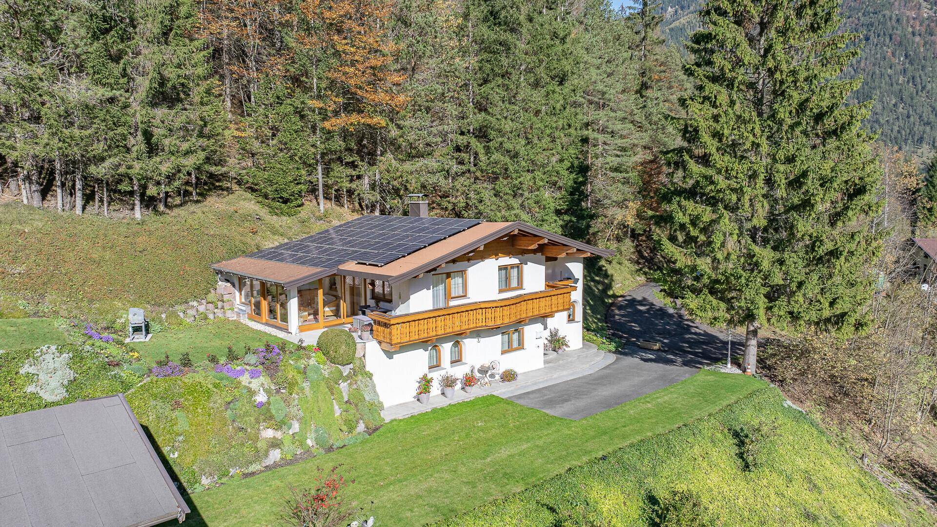 Luftaufnahme eines Alpenchalets mit Photovoltaikanlage auf dem Dach, umgeben von Wald und Bergen.