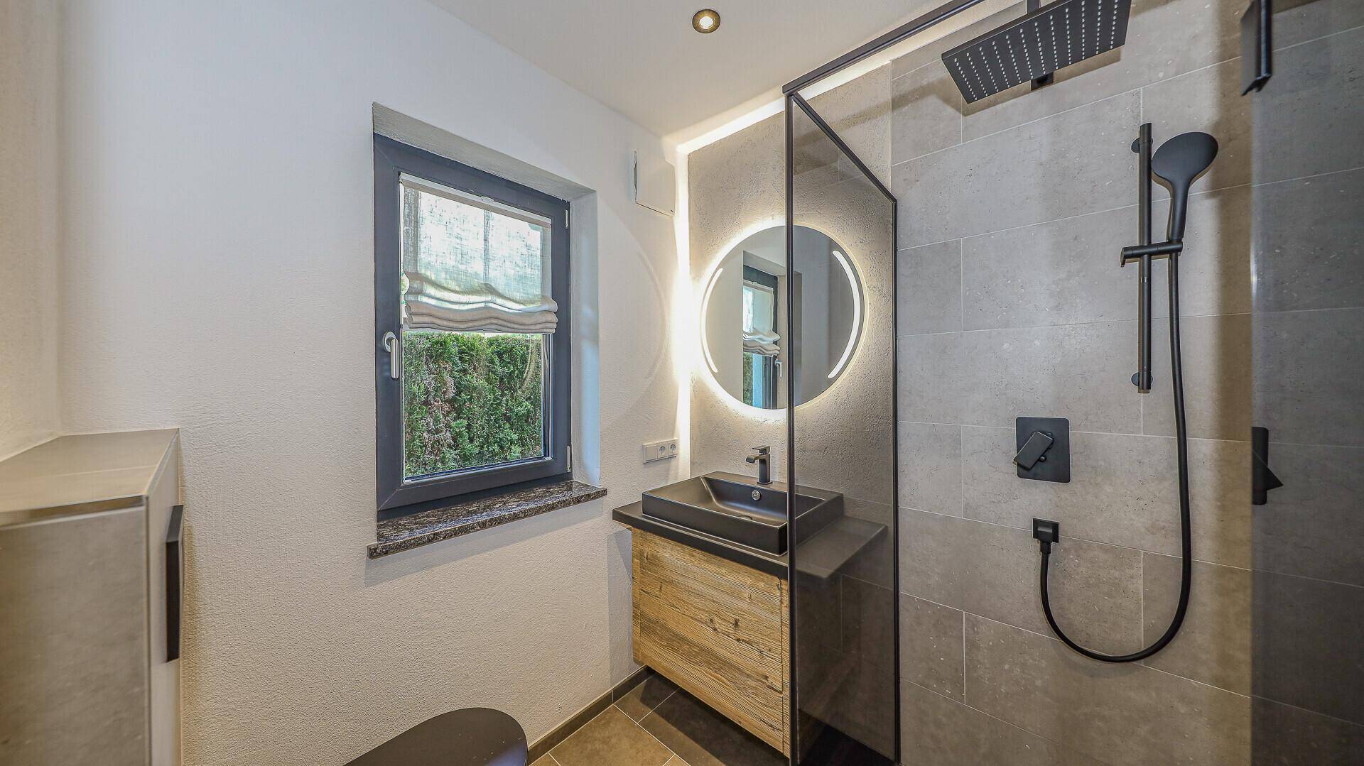 Modernes Badezimmer mit begehbarer Dusche, beleuchtetem Spiegel und Fenster.
