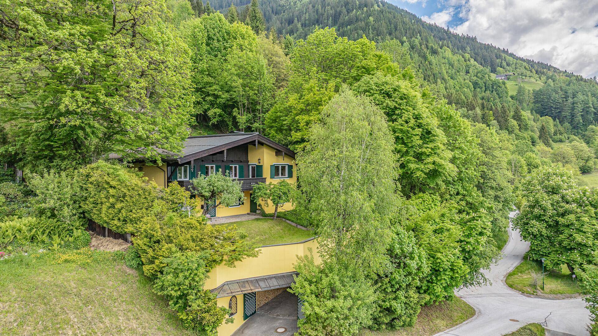 Luftaufnahme des gelben Hauses mit Garten, überdachtem Parkplatz und viel Grün.