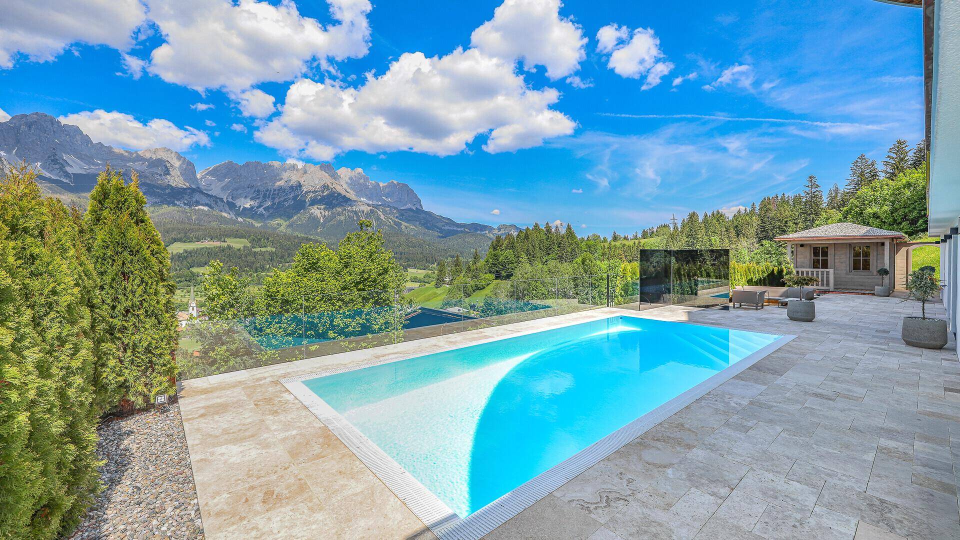 Sonniger Außenpoolbereich mit Terrasse, Liegestühlen und beeindruckendem Bergblick.