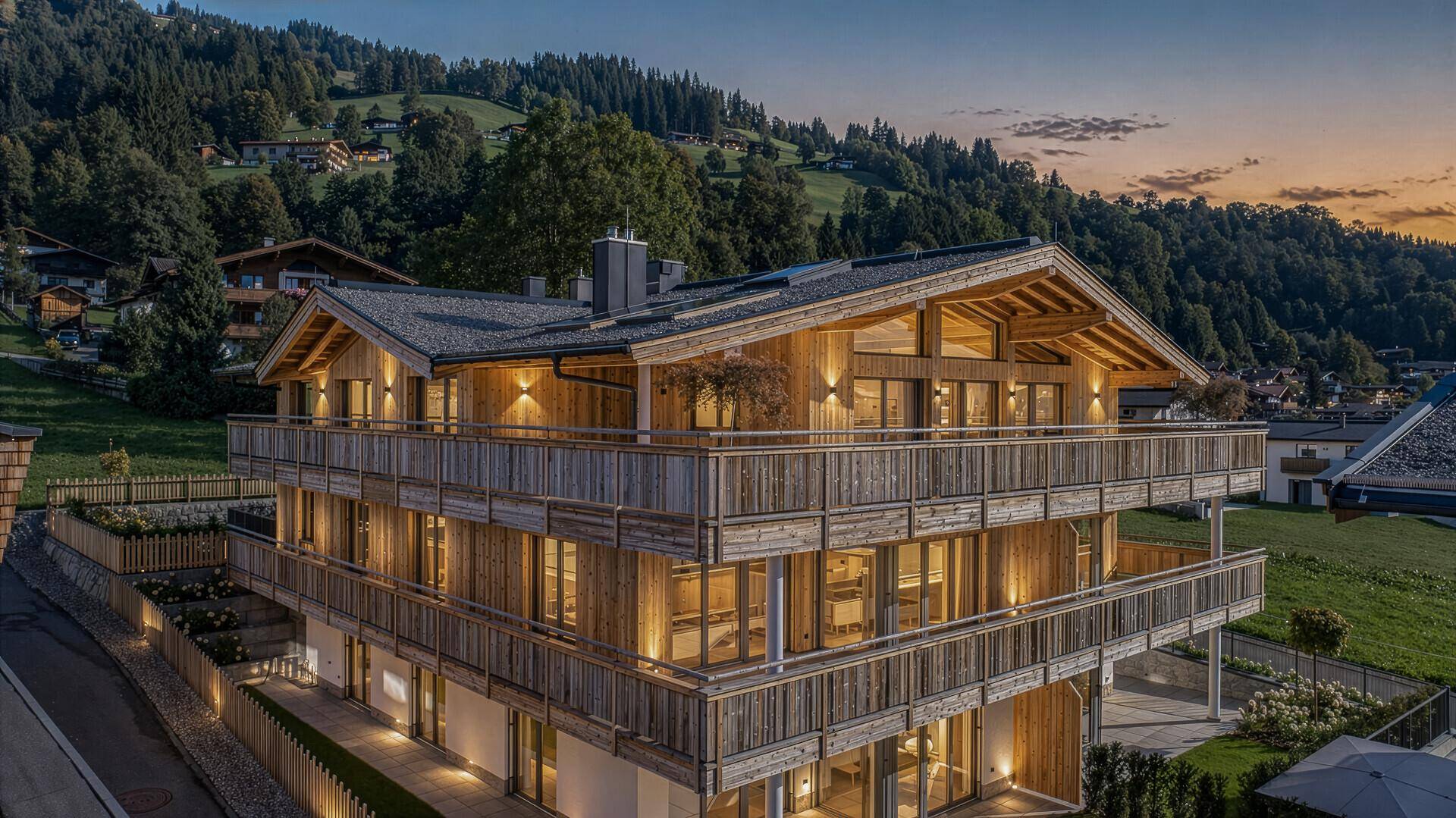 Attraktives Mehrfamilienhaus im alpinen Stil mit Holzbalkonen und Beleuchtung bei Dämmerung.