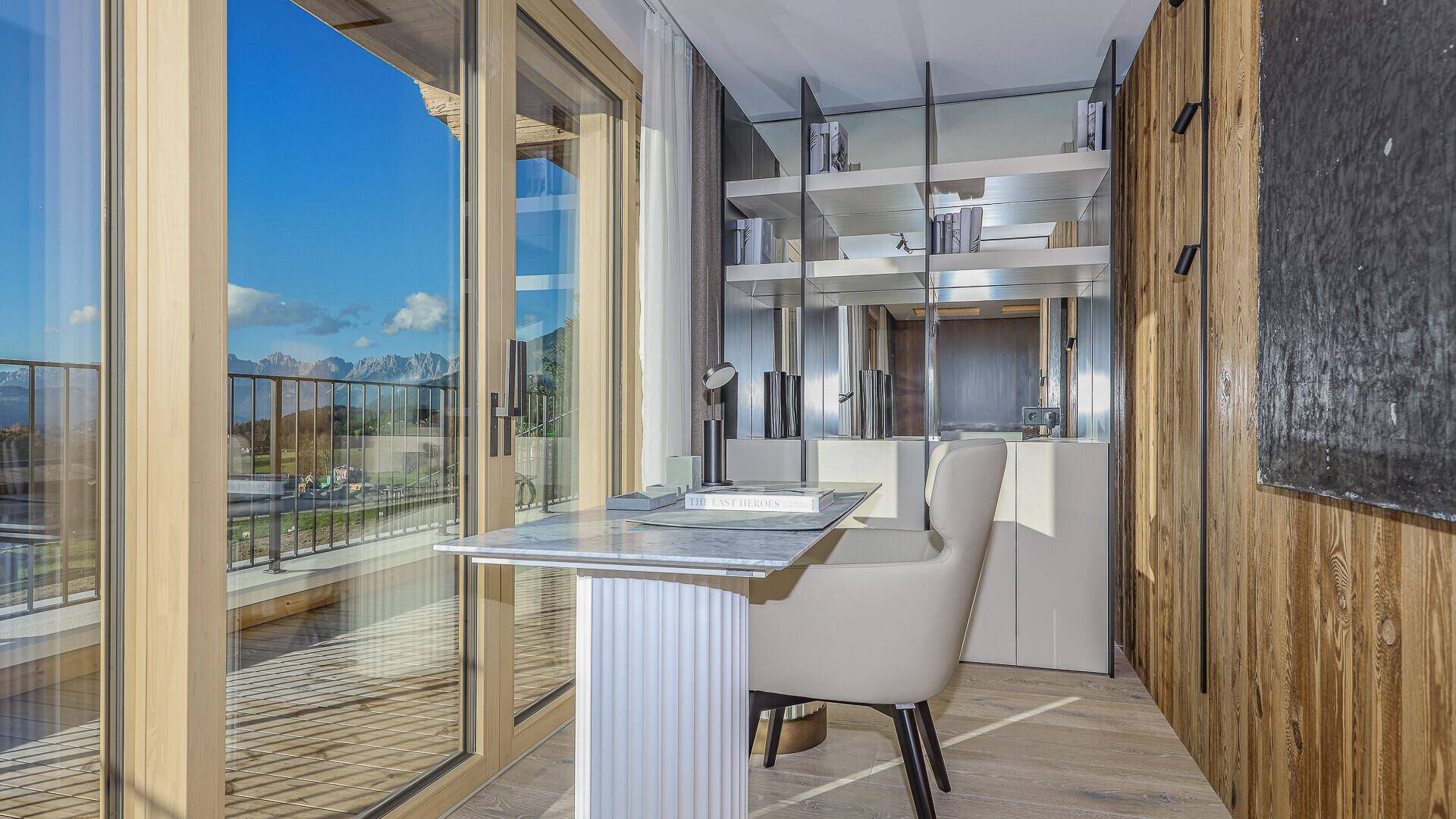 Heller Arbeitsbereich mit Schreibtisch, Bücherregal und Zugang zu einem Balkon mit Bergblick.
