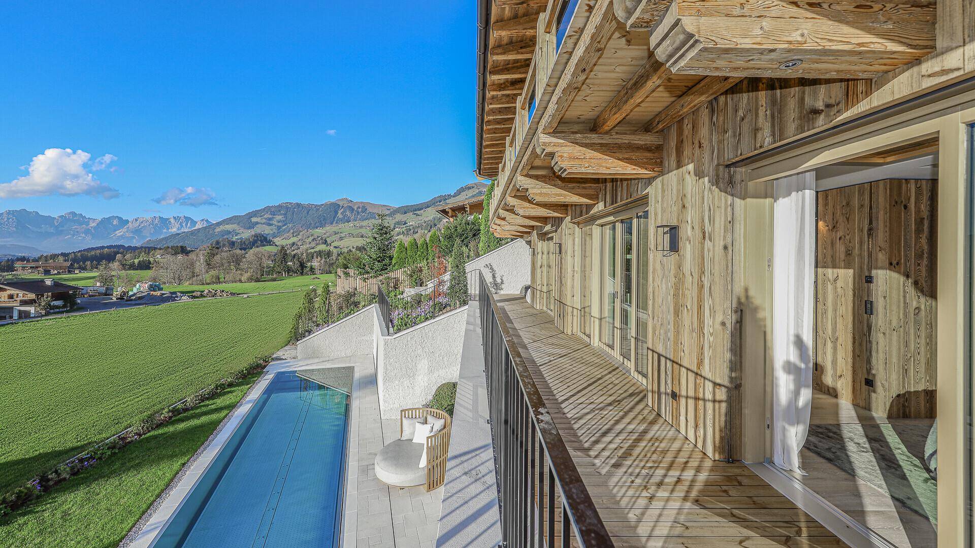 Blick von einem Holz-Balkon auf grüne Felder und eine beeindruckende Berglandschaft unter blauem Himmel.