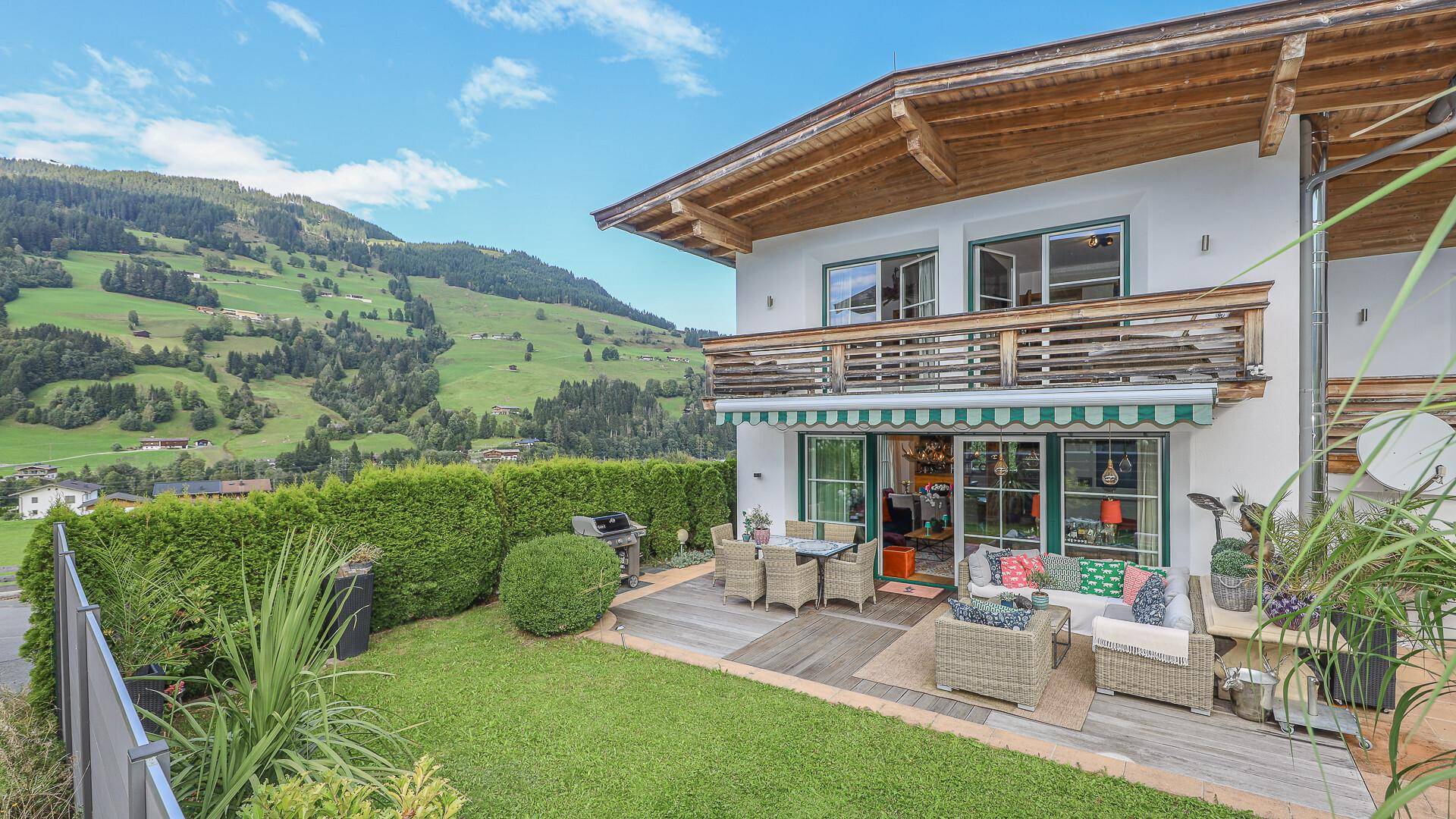 Gepflegter Gartenbereich mit Terrasse, Rasenfläche und Hecke, umgeben von grünen Hügeln.