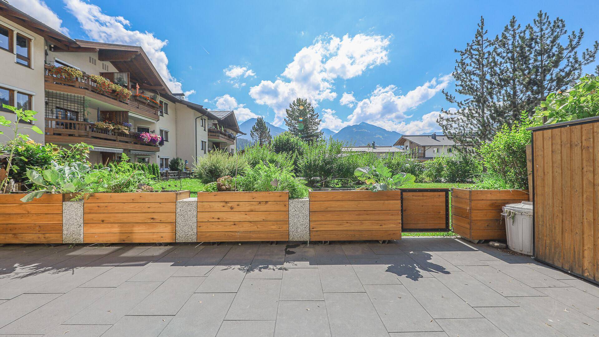 Gepflasterte Terrasse mit erhöhten Gartenbeeten und Blick auf die Berge und umliegende Gebäude.