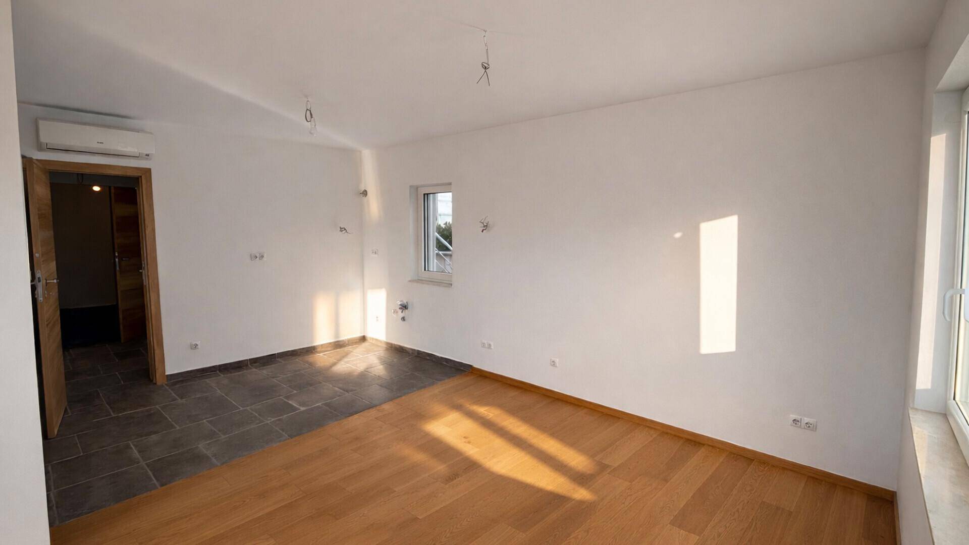 Heller Raum mit Holzboden und Fliesenbereich, ausgestattet mit Klimaanlage und Fenster.