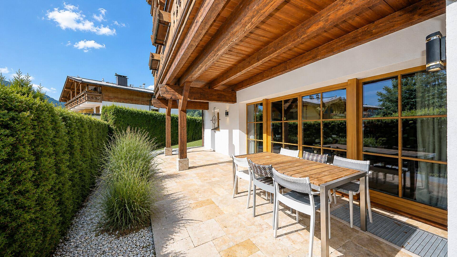 Überdachte Terrasse mit Esstisch und Stühlen, umgeben von Grün und mit Holzbalken.