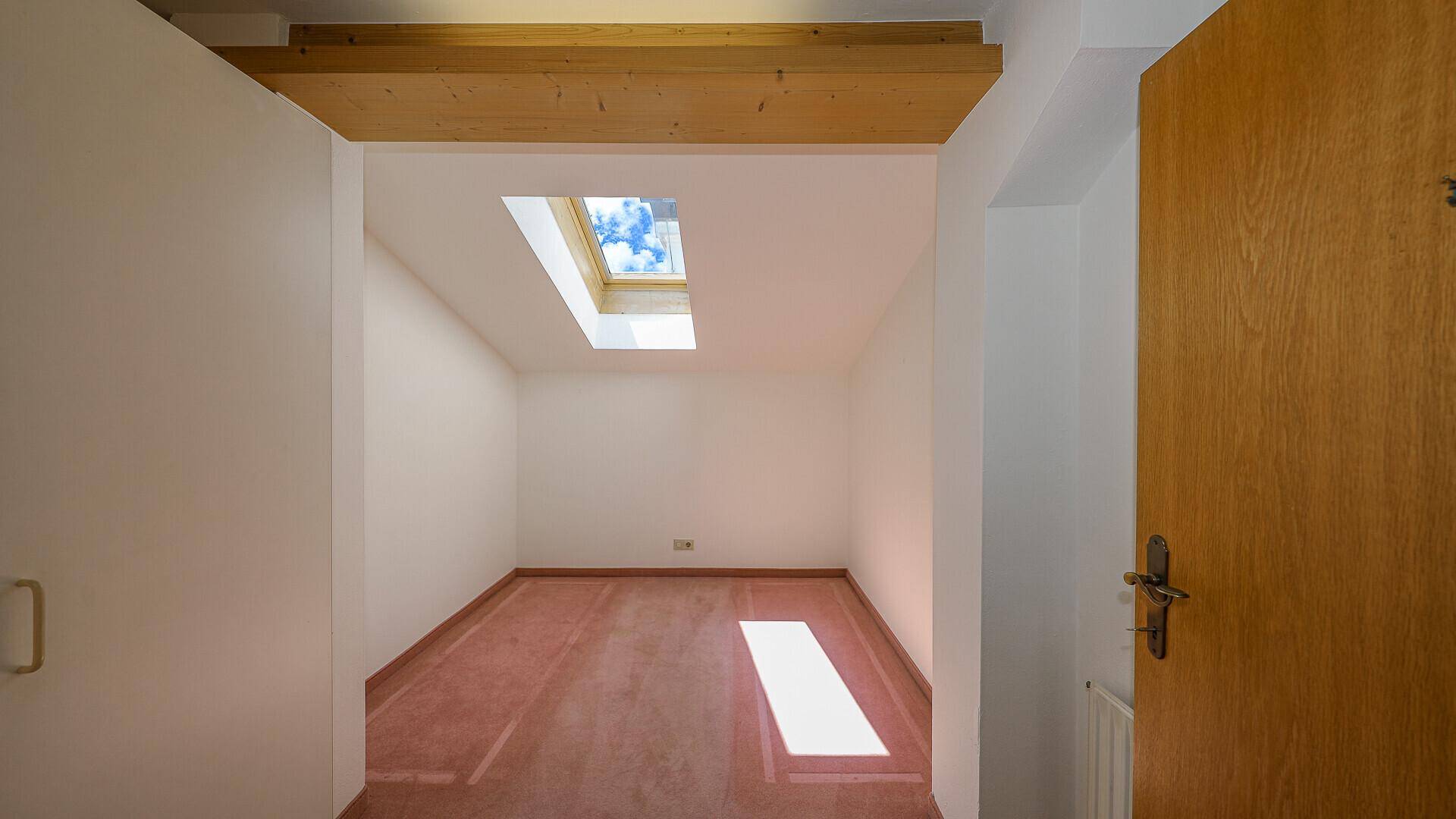 Kleiner, leerer Raum mit Dachfenster, Holzbalken und unfertigem Bodenbelag.