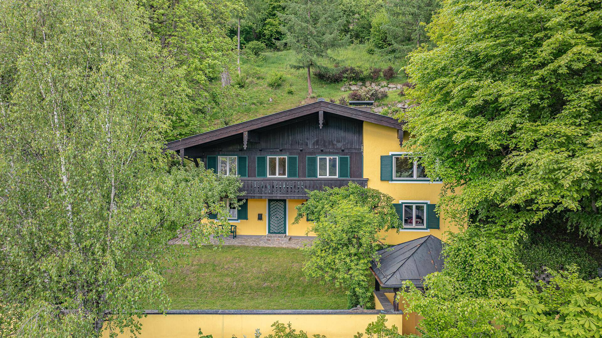 Traditionelles Haus mit gelber Fassade, Holzdetails und grünen Fensterläden in alpiner Umgebung.