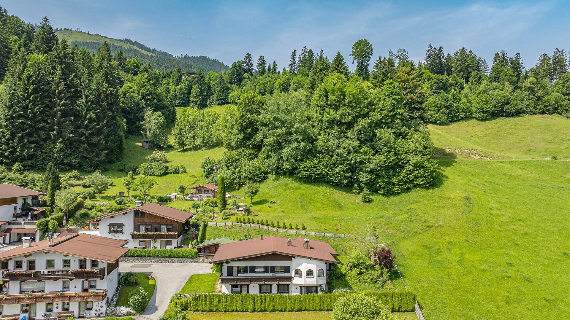 Mehrere Wohnhäuser eingebettet in eine weitläufige grüne Hügellandschaft unter blauem Himmel.