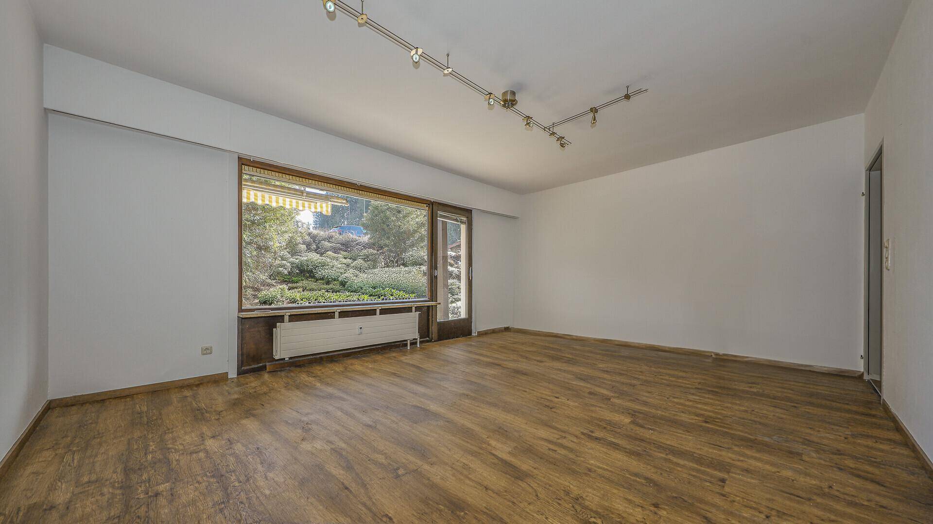 Leerer Wohnraum mit Holzboden, großen Fenstern und Blick auf die grüne Umgebung.