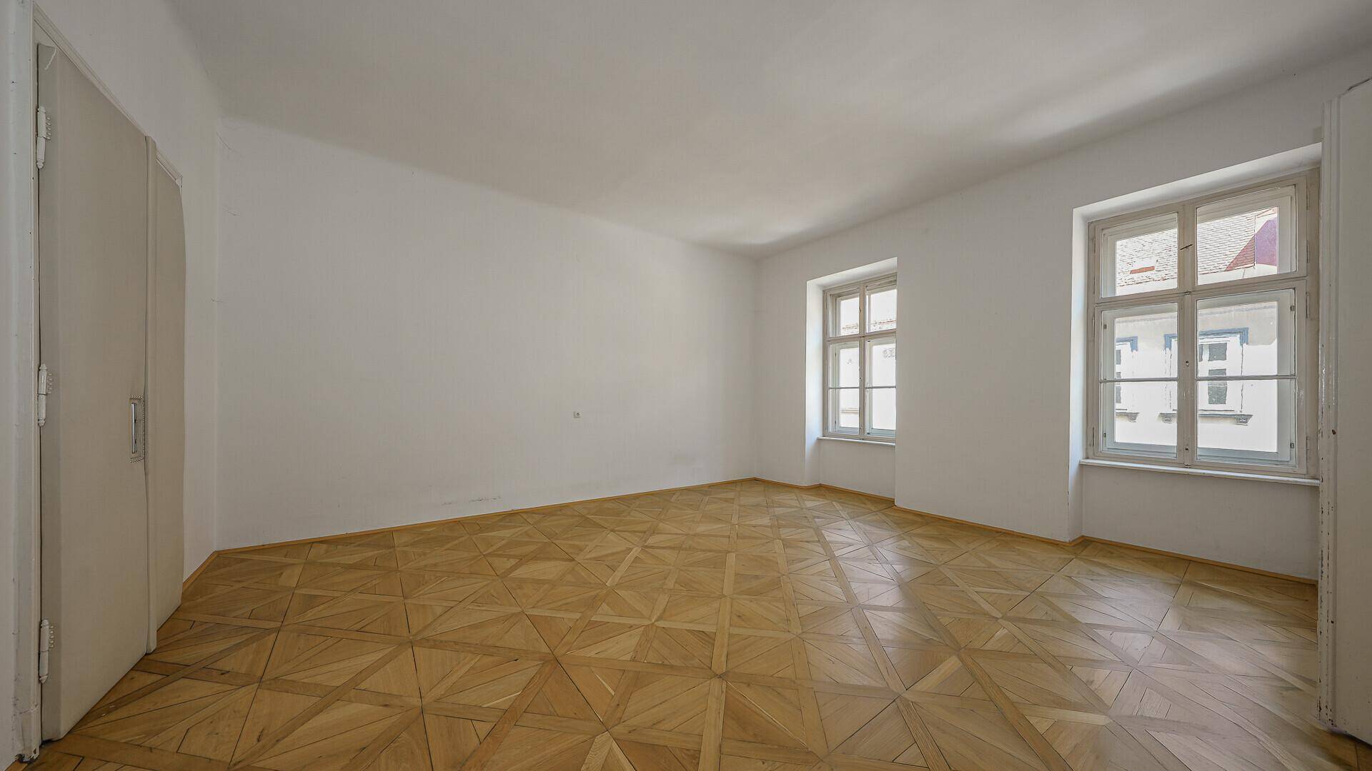 Geräumiges, leeres Zimmer mit hellem Parkettboden und zwei großen Fenstern.