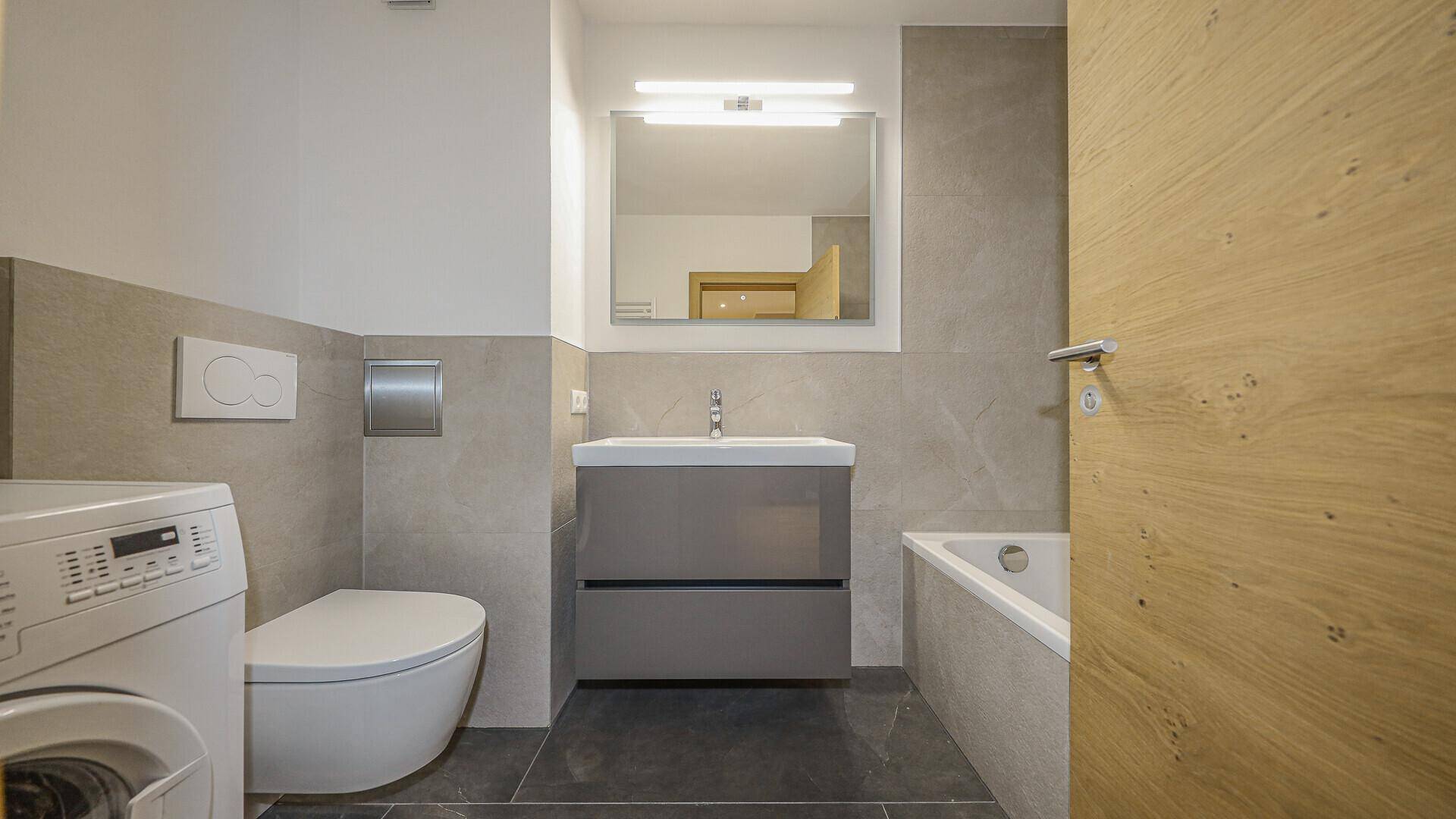 Modernes Badezimmer mit grauen Fliesen, Waschtisch, WC, Badewanne und Waschmaschine.