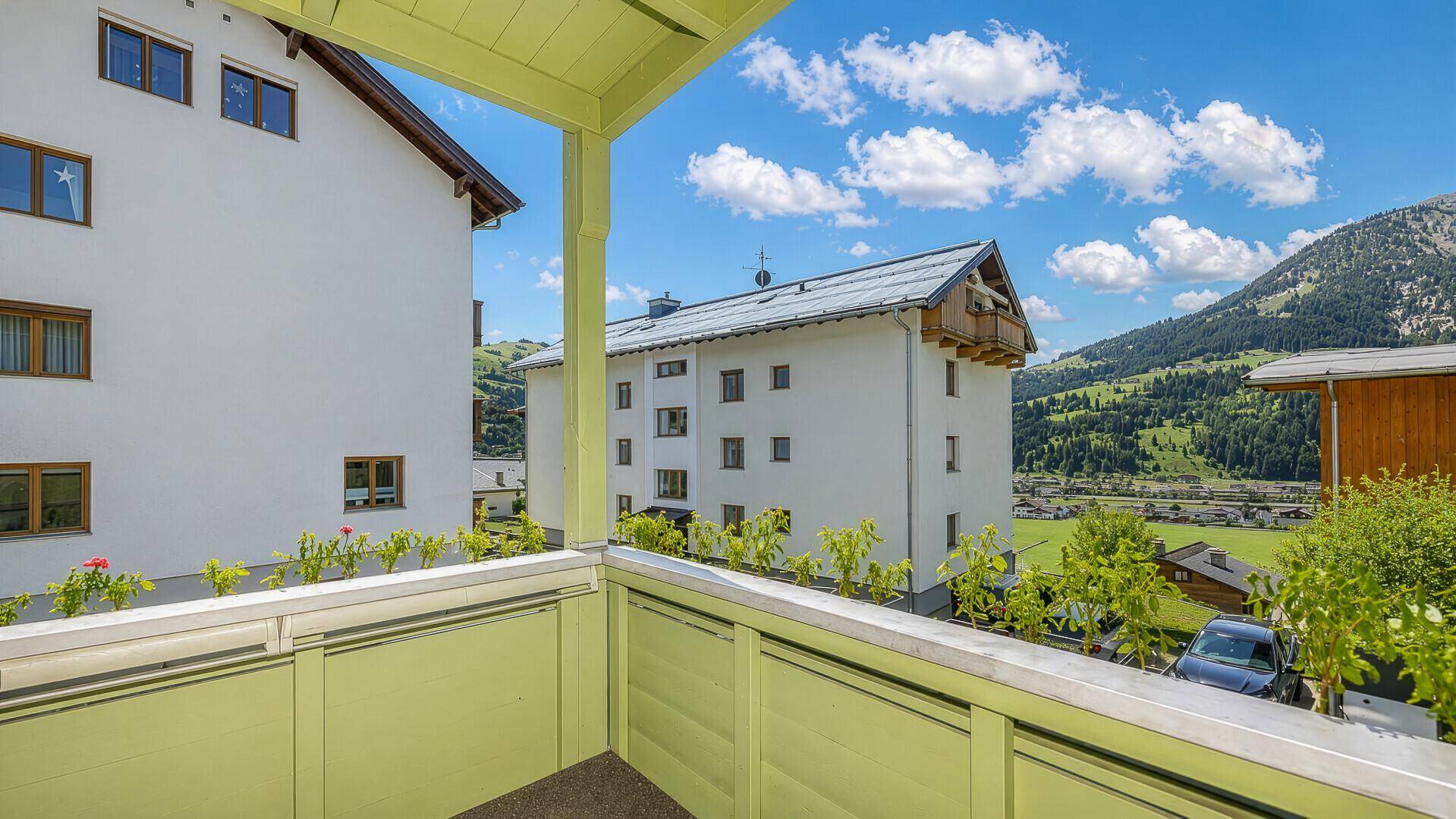 Balkon mit Bergblick und grüner Brüstung, umgeben von alpiner Landschaft und weiteren Gebäuden.