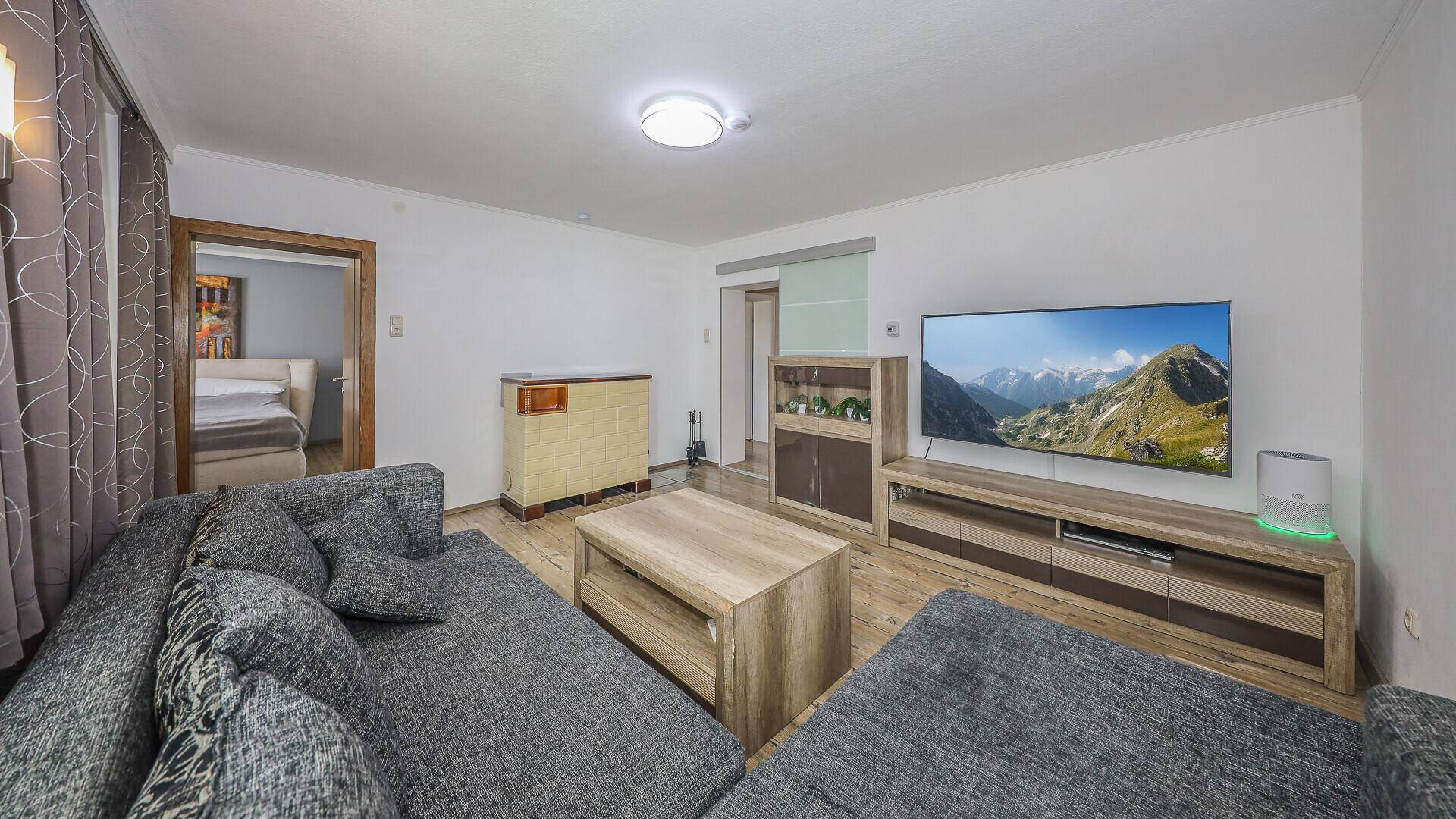 Helles Wohnzimmer mit grauem Ecksofa, Holztisch und Fernseher, mit Zugang zum Schlafzimmer.