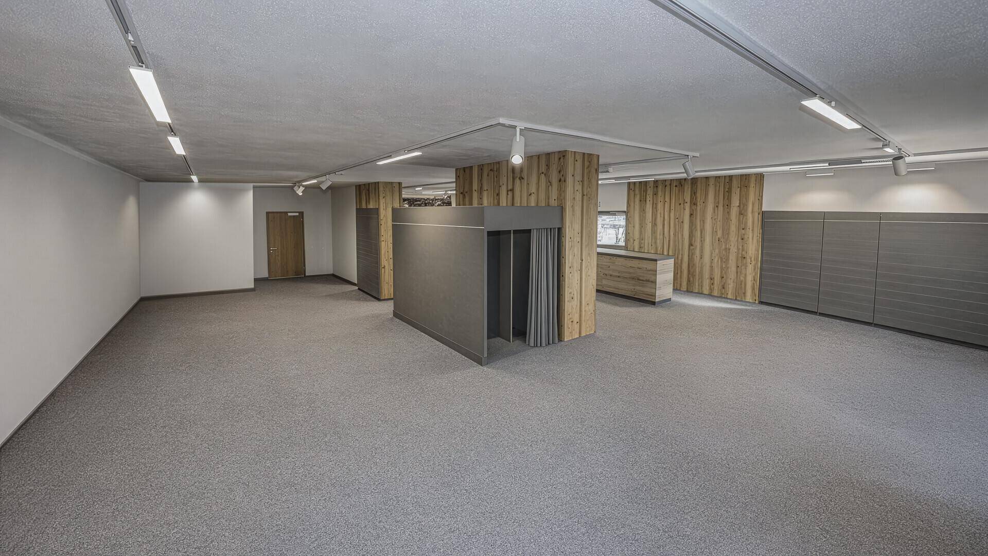 Großer, leerer Gewerberaum mit Teppichboden, Holzwänden und Umkleidekabinen, bereit zur individuellen Gestaltung.
