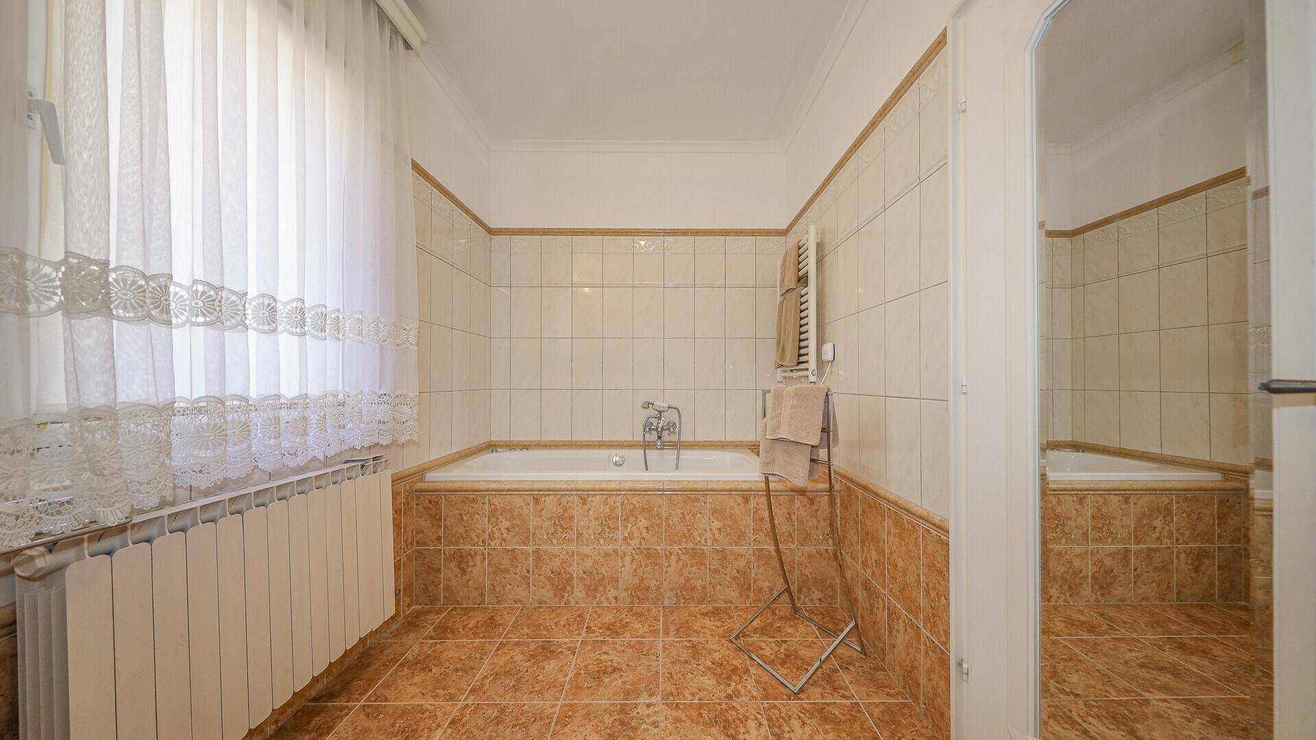 Badezimmer mit Badewanne, beigen Fliesen, beheiztem Handtuchhalter und Fenster für Tageslicht.