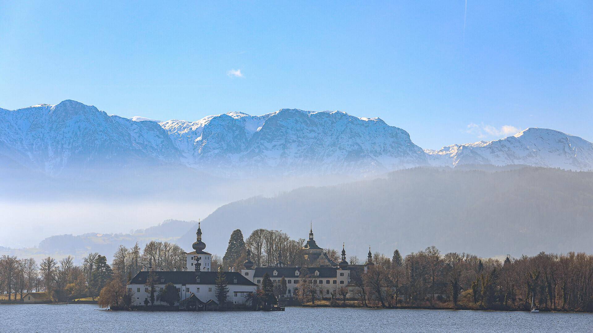 Malerischer Blick auf ein Schloss am Seeufer mit schneebedeckten Bergen im Hintergrund.