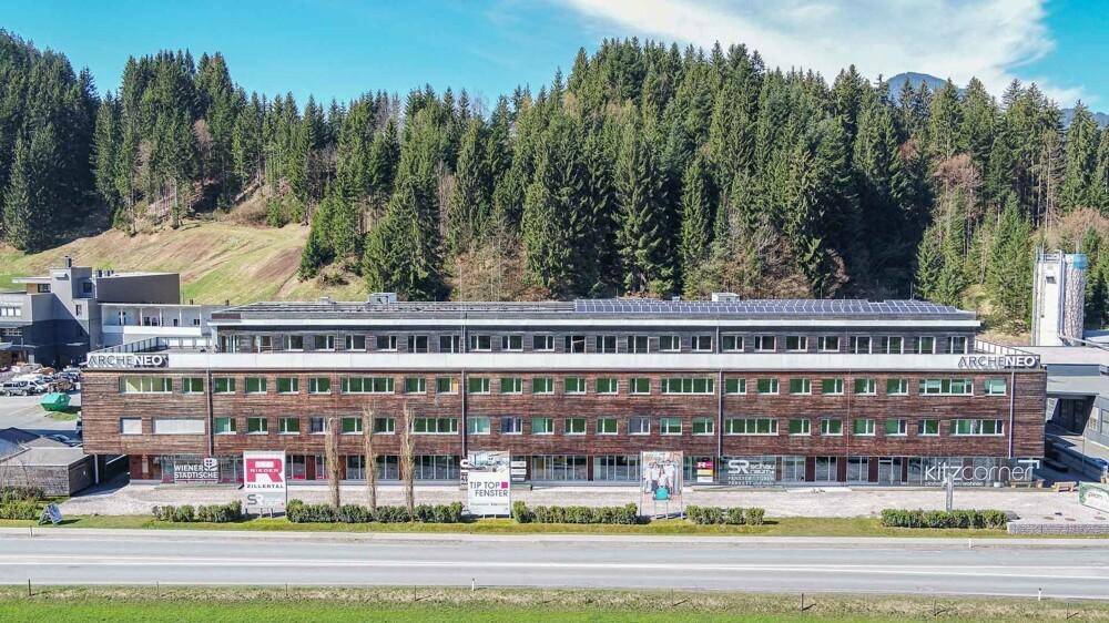 Frontale Luftaufnahme eines modernen Gebäudes mit Holzfassade, umgeben von Wald und einer Straße.