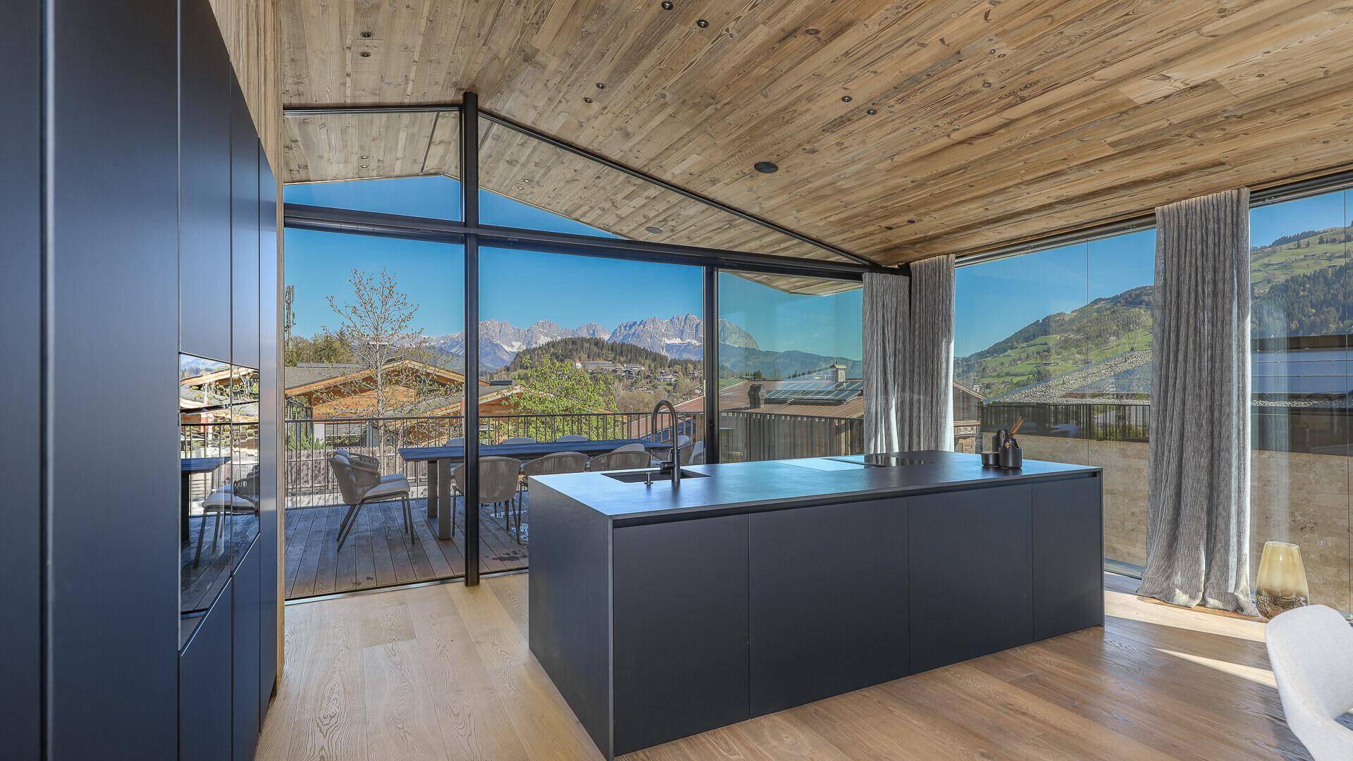 Offene, moderne Küche mit dunklen Fronten, Kücheninsel und direktem Zugang zur Terrasse mit Bergblick.