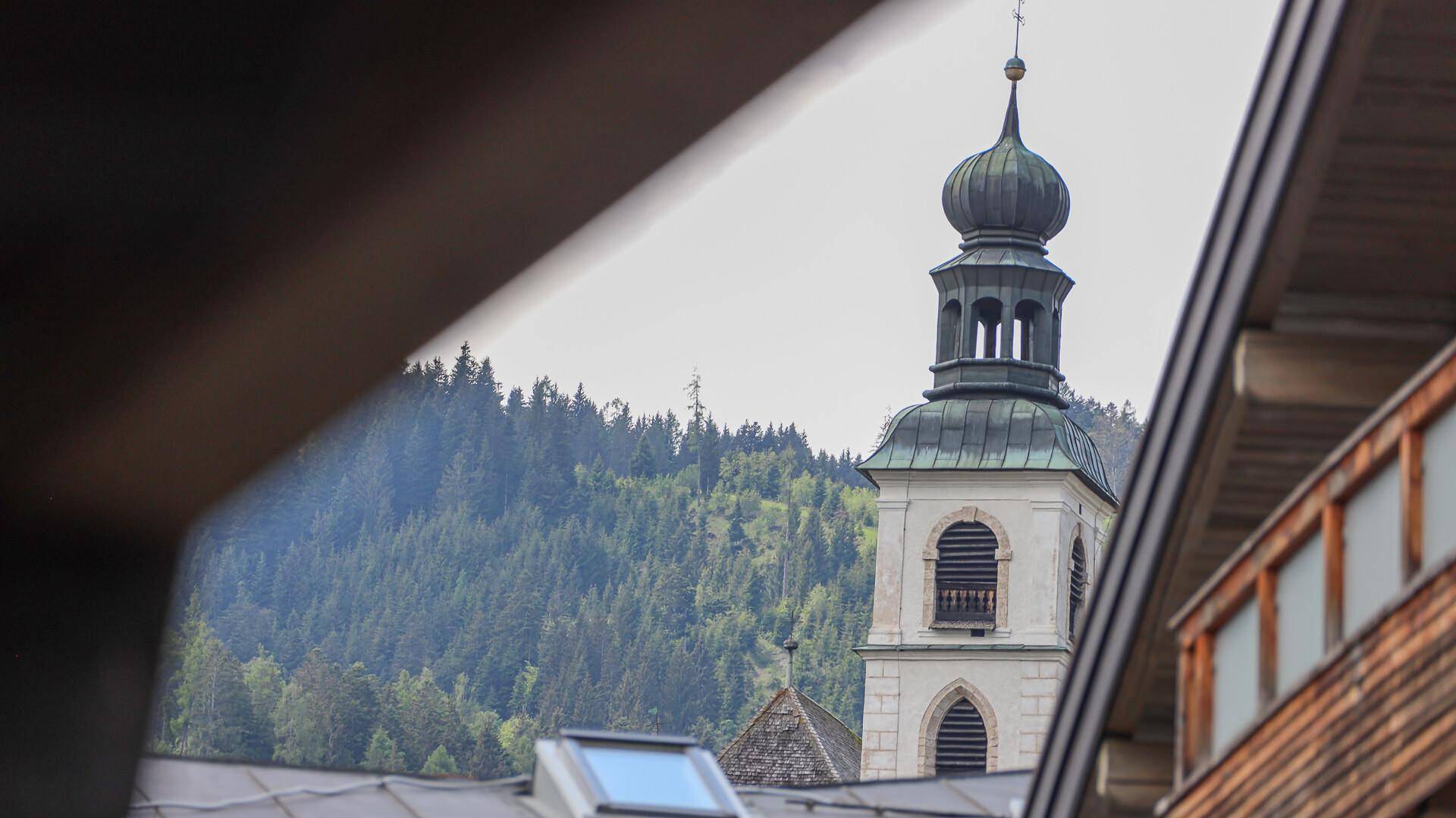 Historischer Kirchturm mit grüner Kuppel, umgeben von bewaldeten Hügeln, durch Dachschrägen gerahmt.