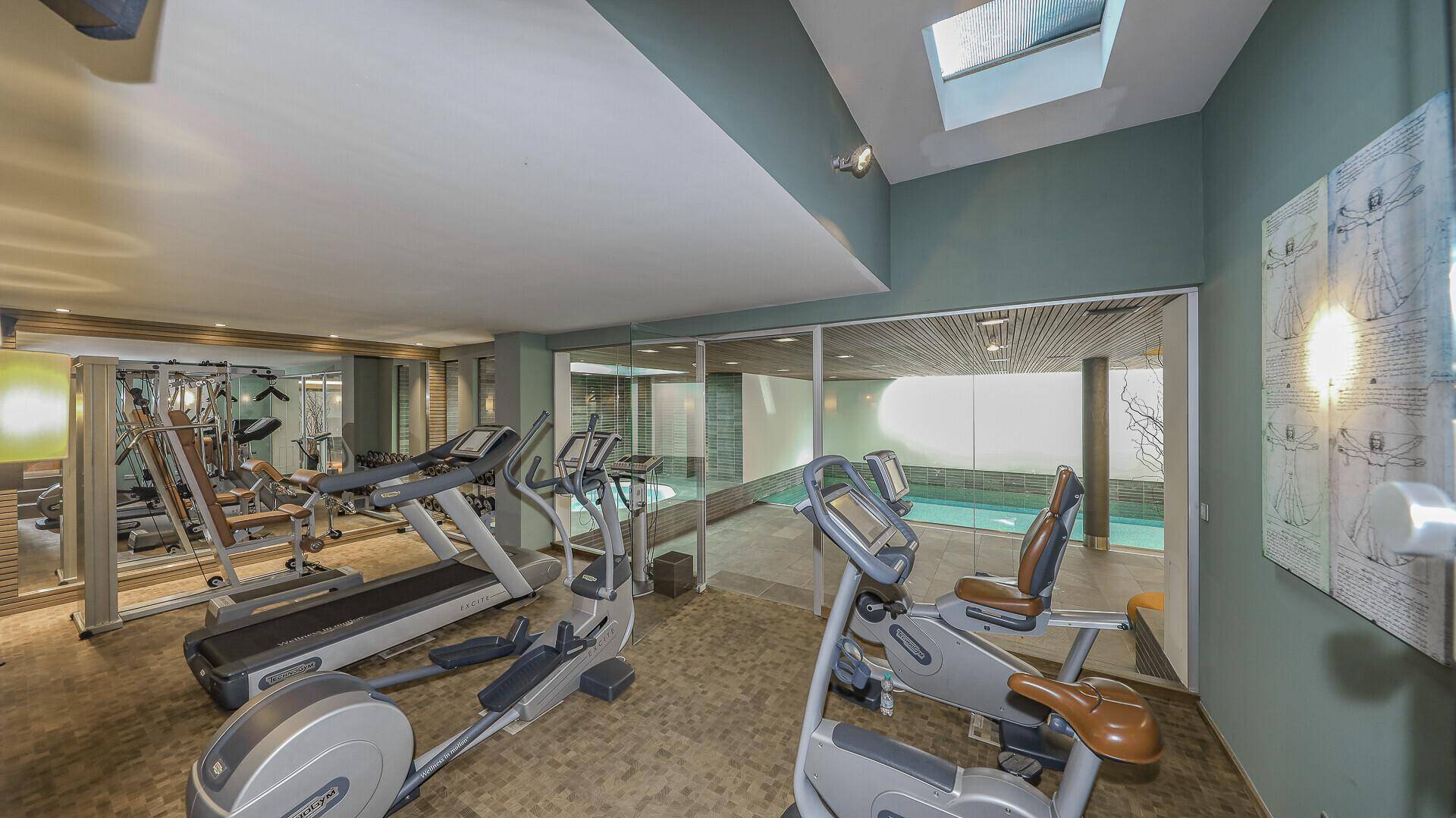 Gut ausgestatteter Fitnessraum mit verschiedenen Trainingsgeräten und Blick auf den Poolbereich.