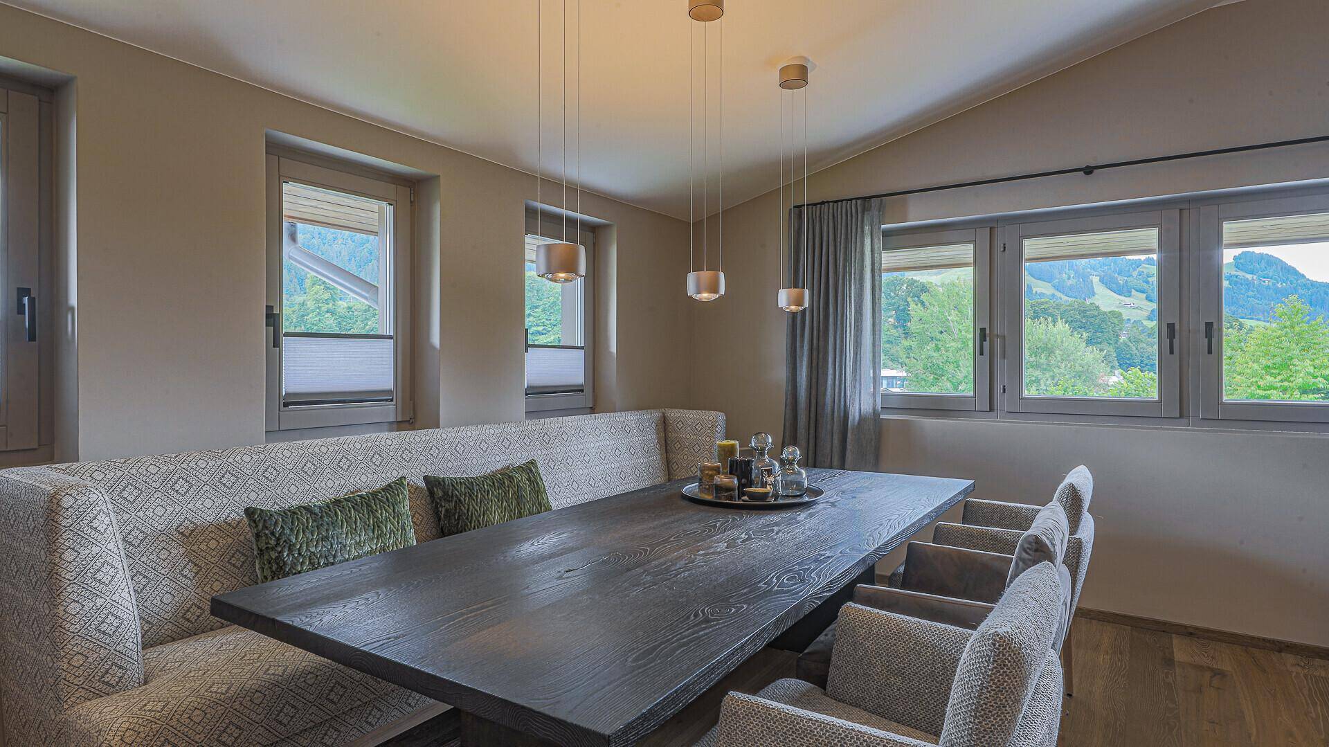 Eleganter Essbereich mit großem Holztisch, bequemer Sitzbank und Fenstern mit grüner Aussicht.